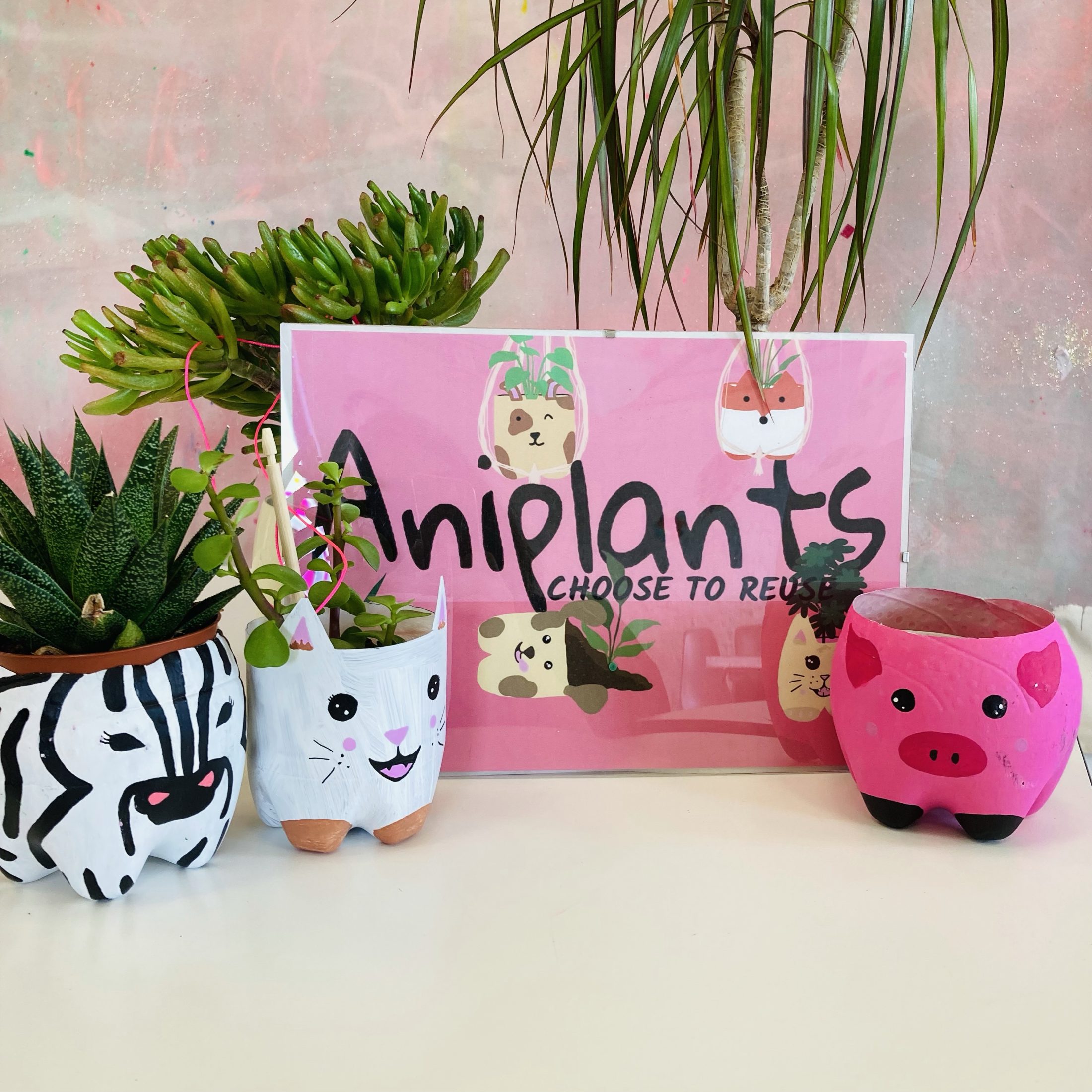 Three animal-themed plant pots (zebra, rabbit, pig) made from reused materials sit next to a pink Aniplants: Choose to Reuse sign, with green leafy plants in the background.
