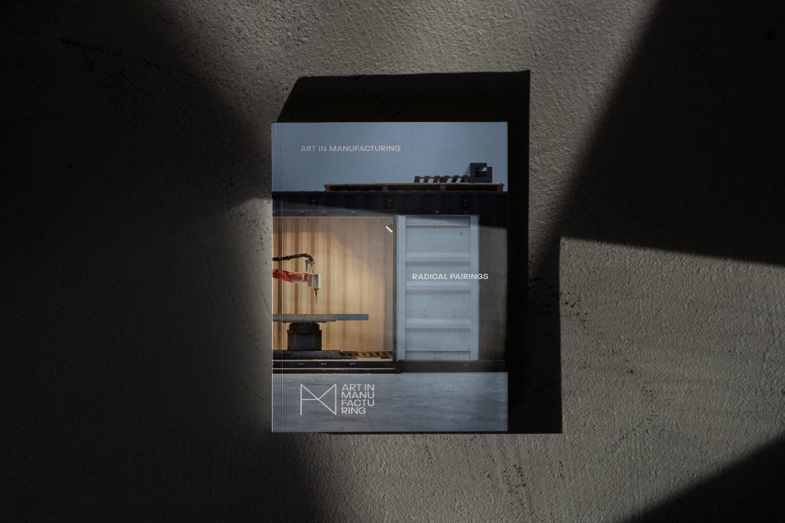 A booklet titled Art in Manufacturing: Radical Pairings lies on a grey, textured surface with dramatic shadows cast across it. The cover shows industrial equipment and a modern interior design.