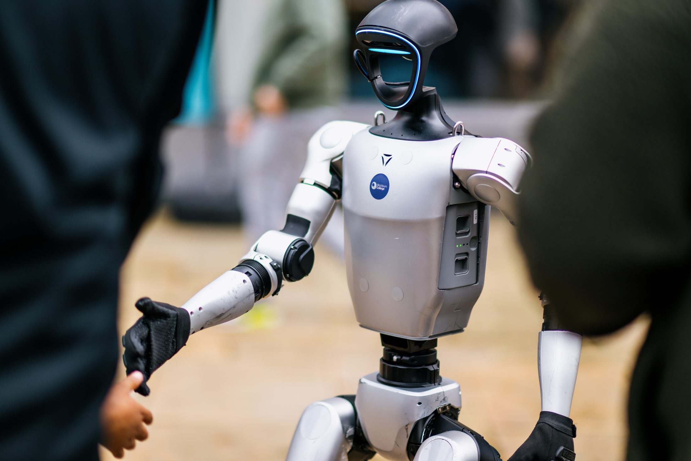 A humanoid robot with a sleek, silver body and a dark visor shakes hands with a person in an outdoor setting, surrounded by blurred people in the background.