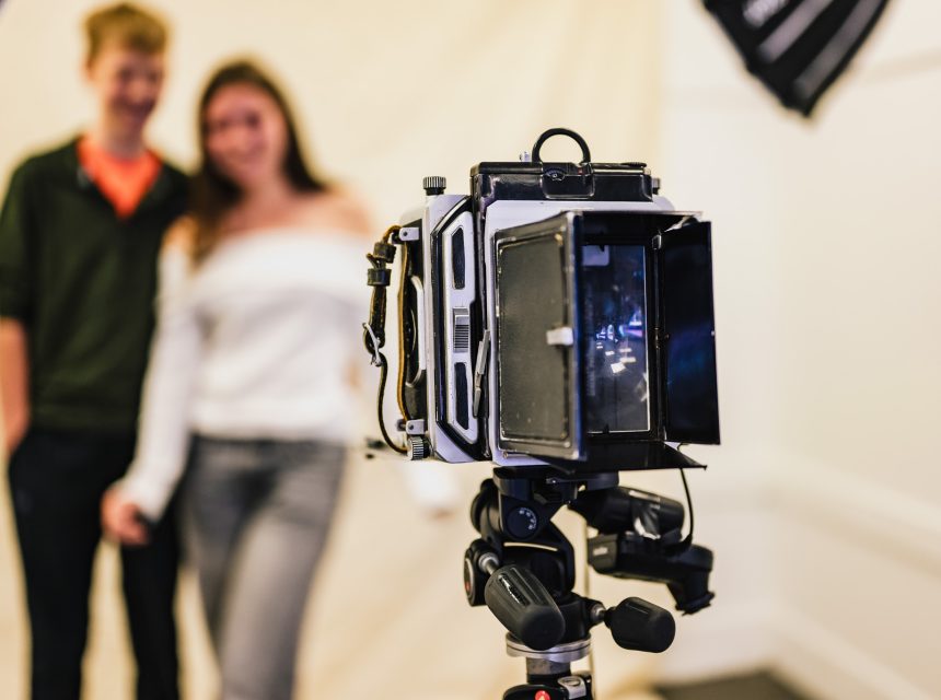 A vintage camera on a tripod is in sharp focus in the foreground, whilst a smiling couple stands blurred in the background against a plain backdrop under studio lights.