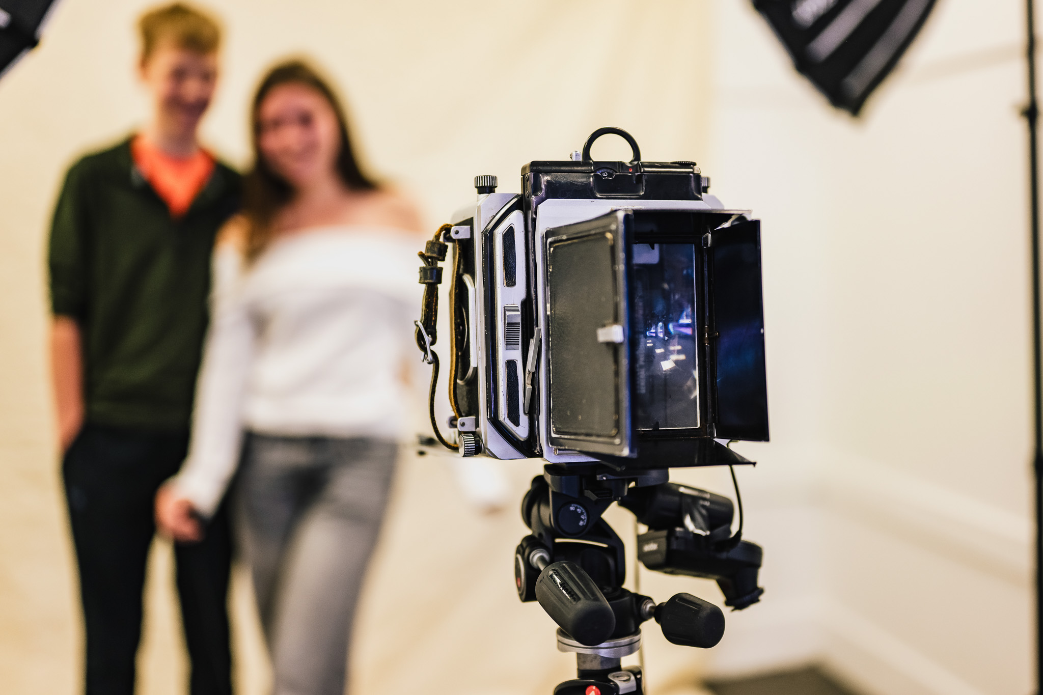 A vintage camera on a tripod is in sharp focus in the foreground, whilst a smiling couple stands blurred in the background against a plain backdrop under studio lights.