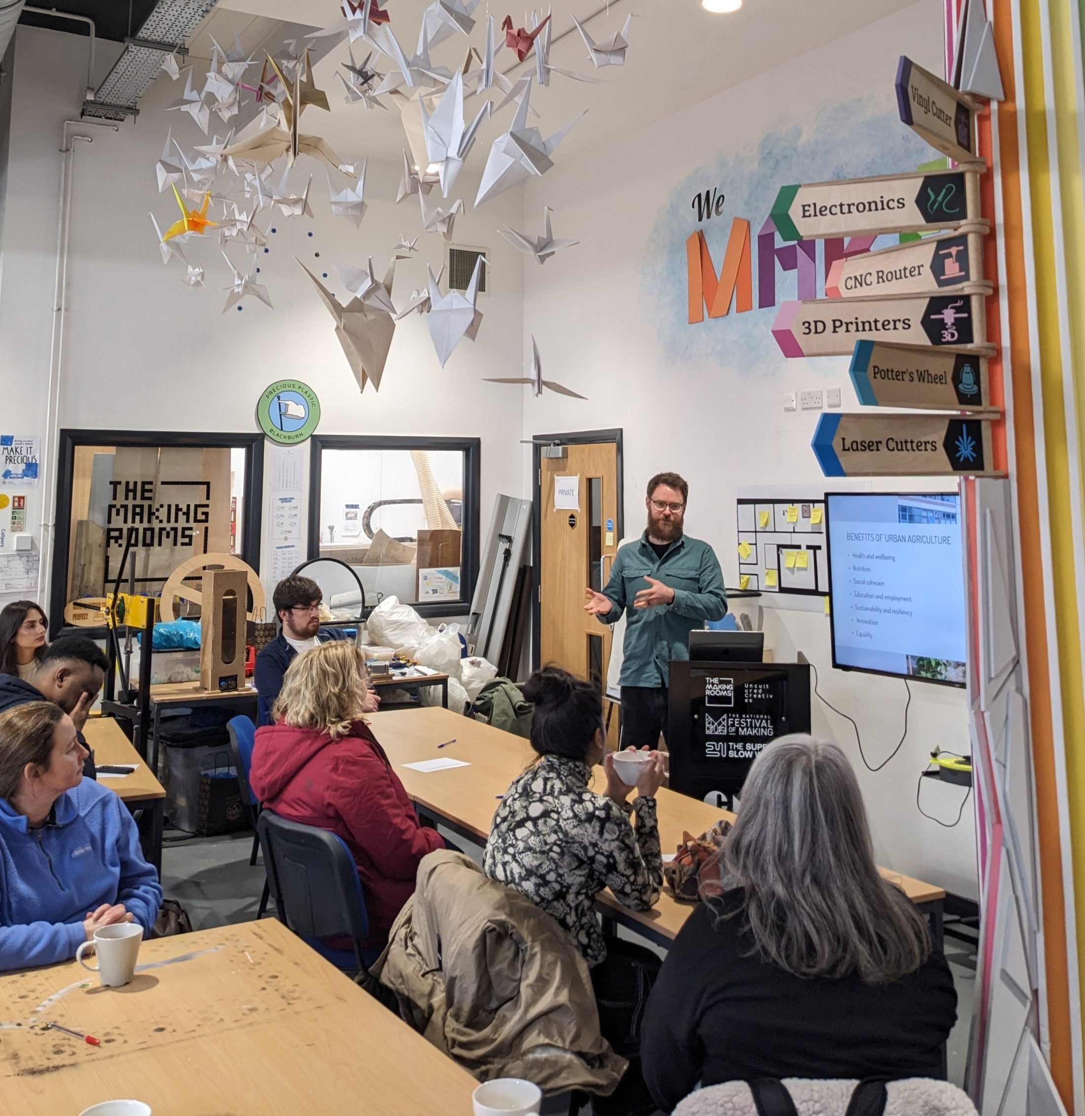A man stands giving a presentation to a small group seated at tables in a makerspace, with colourful origami cranes hanging from the ceiling and signs pointing to various equipment like 3D printers, pottery, and laser cutters.