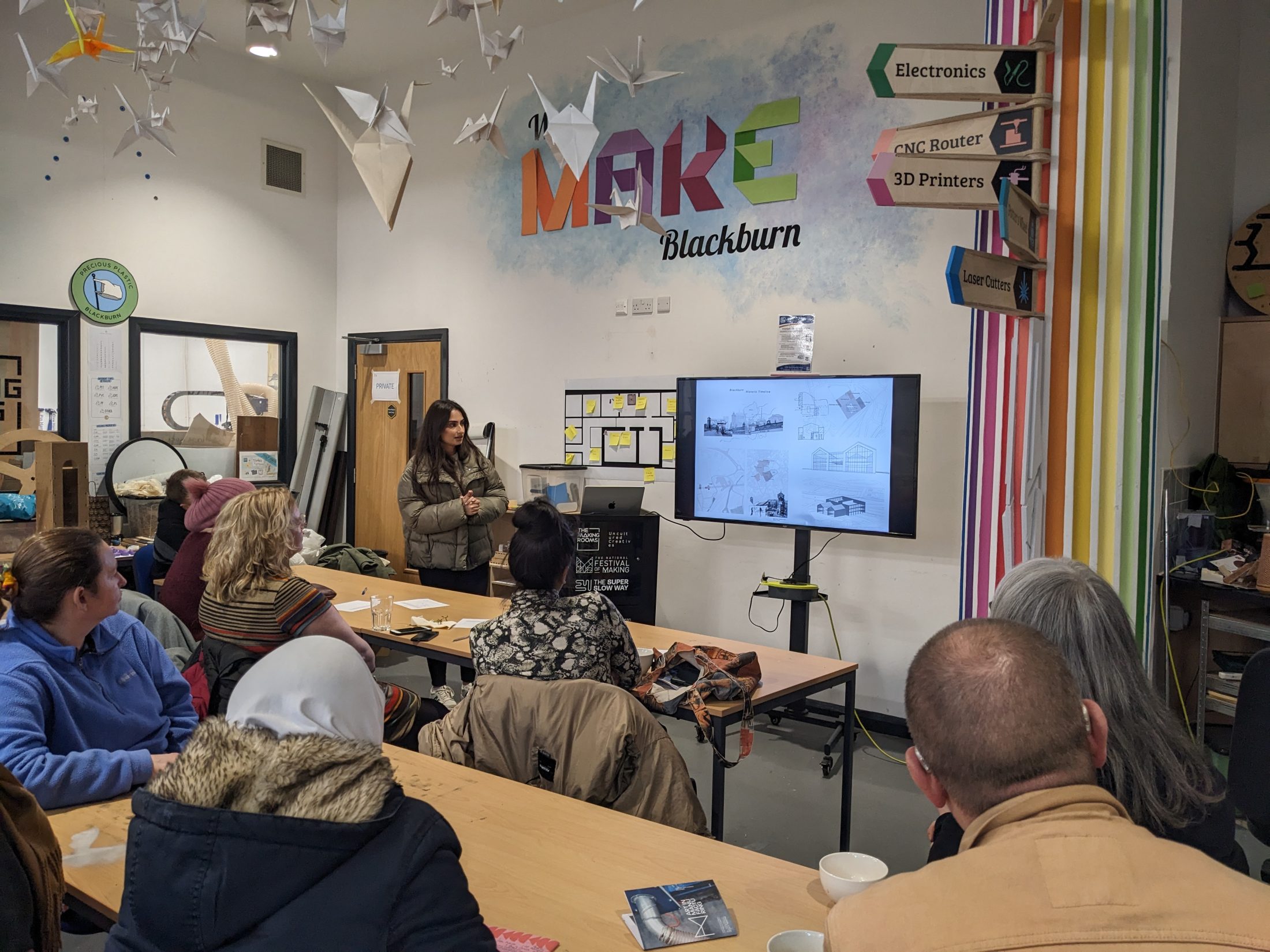 A woman presents architectural plans on a screen to a seated audience in a creative workshop space decorated with paper cranes and colourful signs reading MAKE Blackburn.