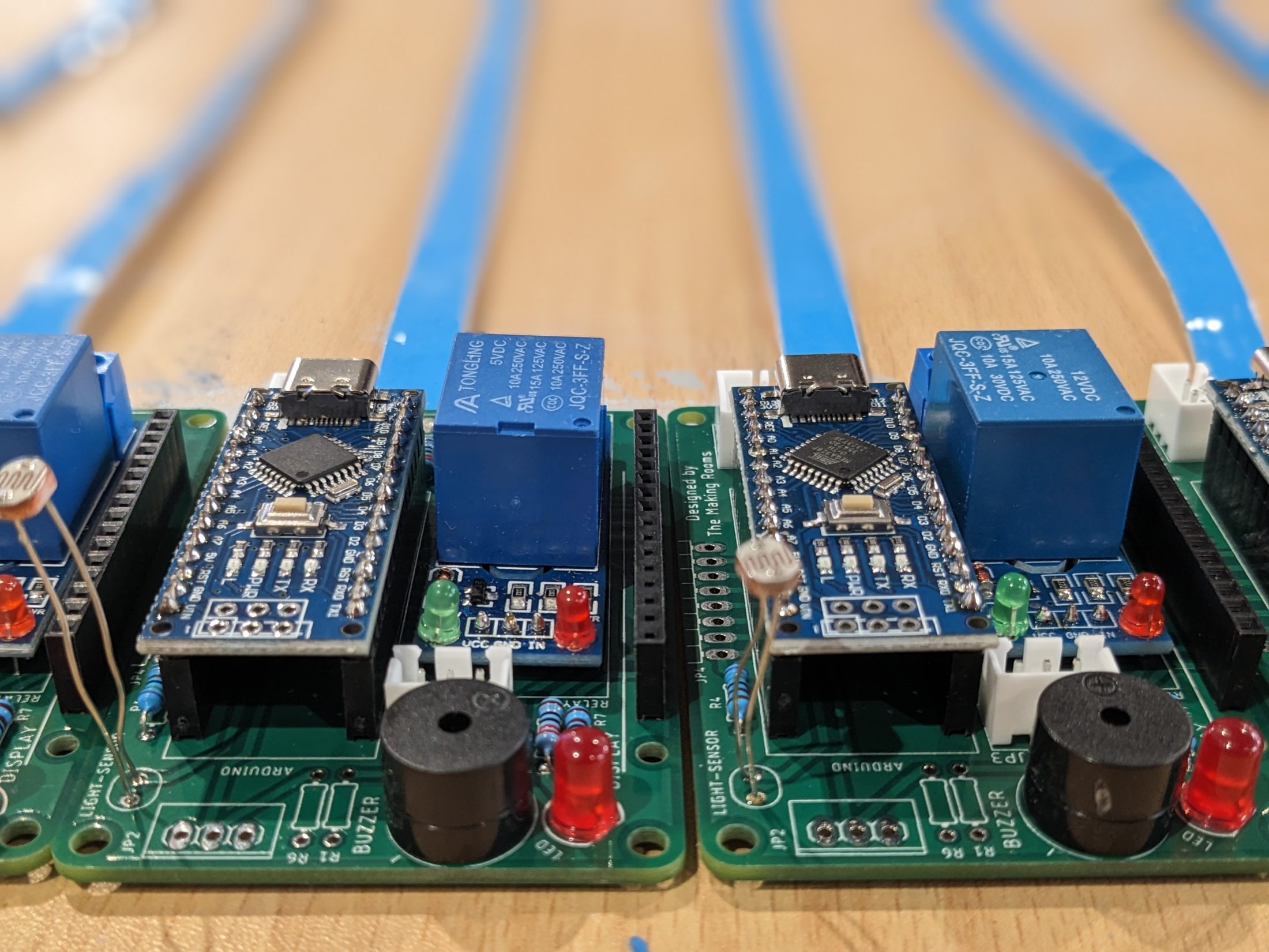 Close-up of electronic circuit boards with blue relays, microcontrollers, red LEDs, and various components arranged in rows on a wooden surface with blue strips in the background.