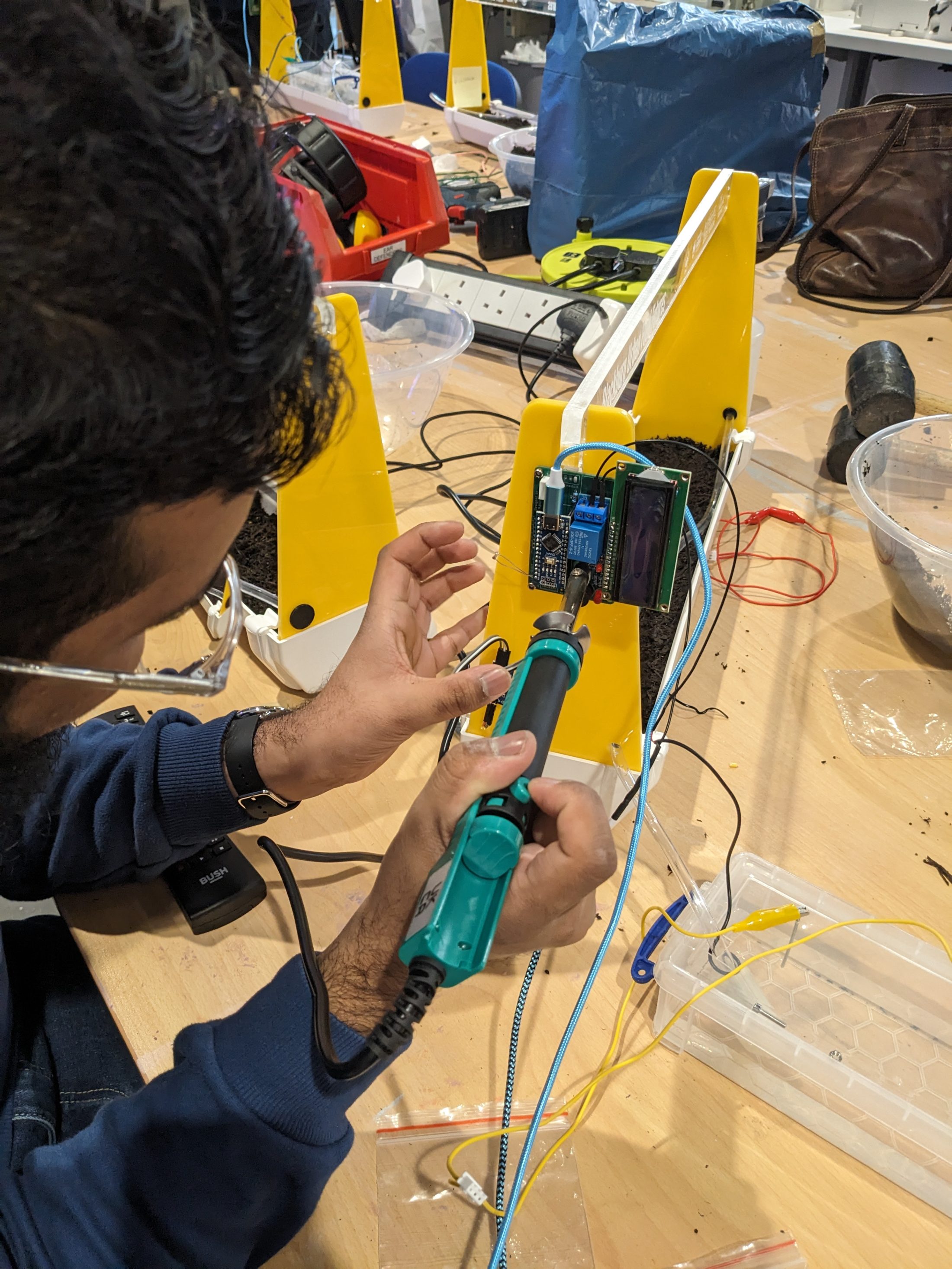 A person uses a soldering iron to work on an electronic circuit attached to a yellow plastic structure on a cluttered workbench with various tools and wires.