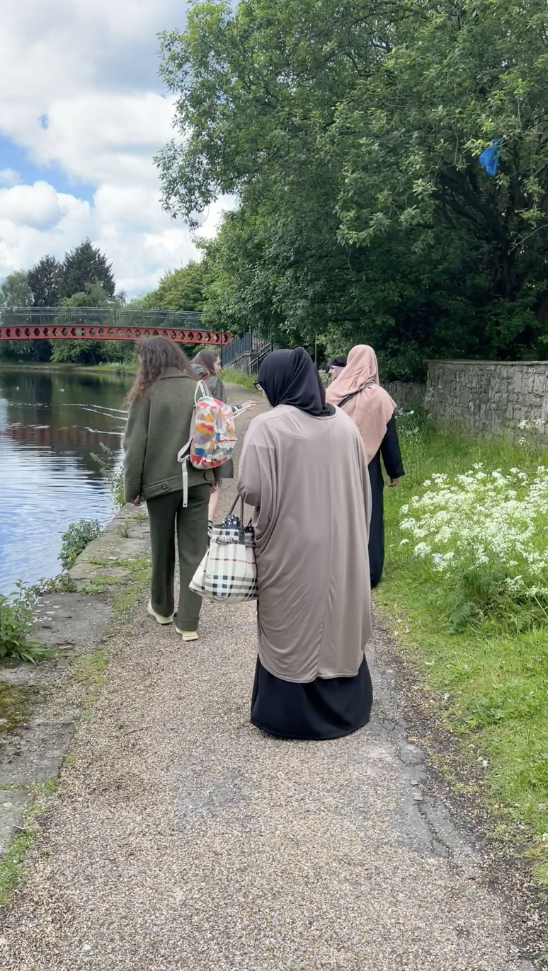 Four women walk along a riverside path bordered by grass and wildflowers, with trees and a red bridge visible in the background. The sky is partly cloudy, and one woman carries a patterned bag.