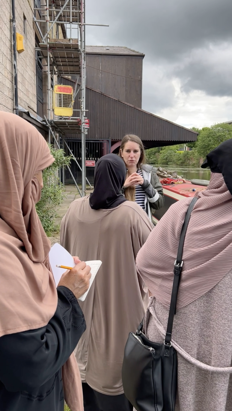A group of women, some wearing hijabs, stand outdoors listening to a woman speaking. One woman takes notes with a pencil and notebook. There is a canal, a red boat, and scaffolding on a nearby building in the background.