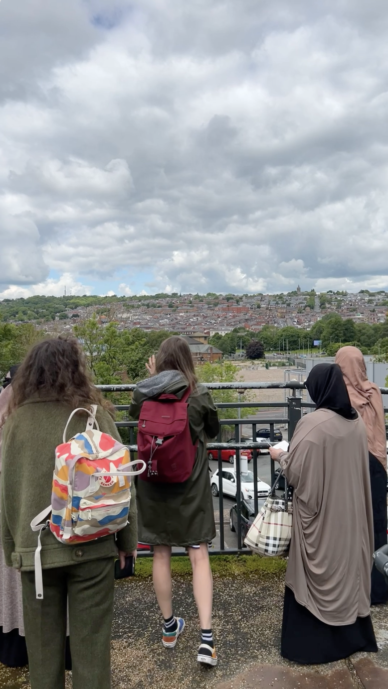 A group of five people with rucksacks and bags stand by a black railing, facing a cityscape with many houses under a cloudy sky. Green trees and parked cars are visible in the background.