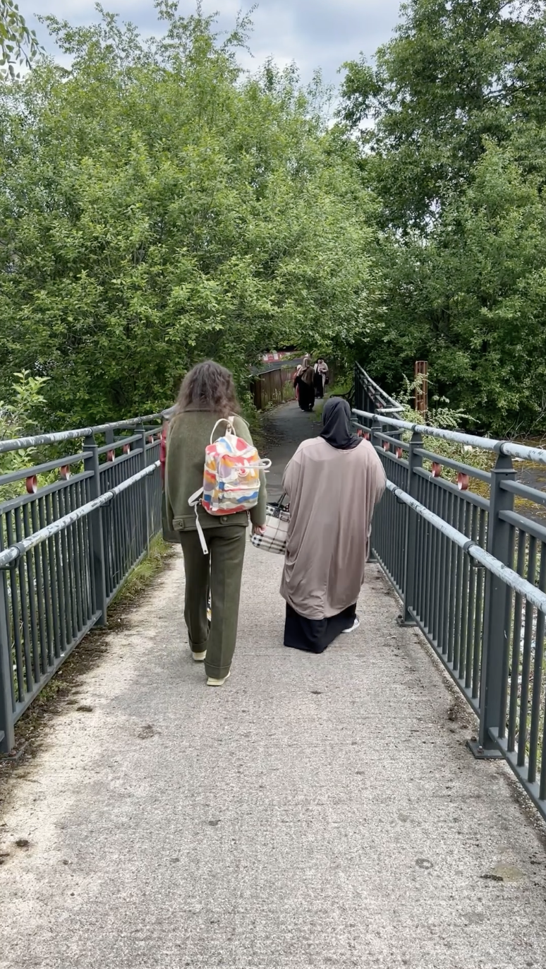 Two people walk across a bridge surrounded by greenery. One carries a colourful rucksack and the other, wearing a headscarf, pushes a shopping trolley. More people can be seen further along the bridge.