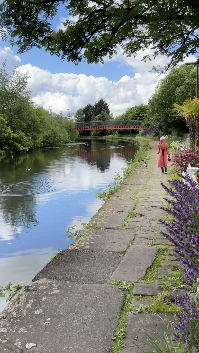 A person in a pink coat stands on a stone path beside a calm canal, with lush greenery, purple flowers, and a red bridge in the background under a partly cloudy sky.