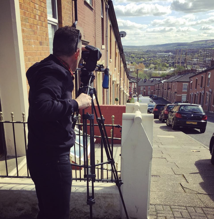 A person wearing a black hoodie operates a video camera on a tripod, filming down a sloping residential street lined with brick houses and parked cars on a partly cloudy day.