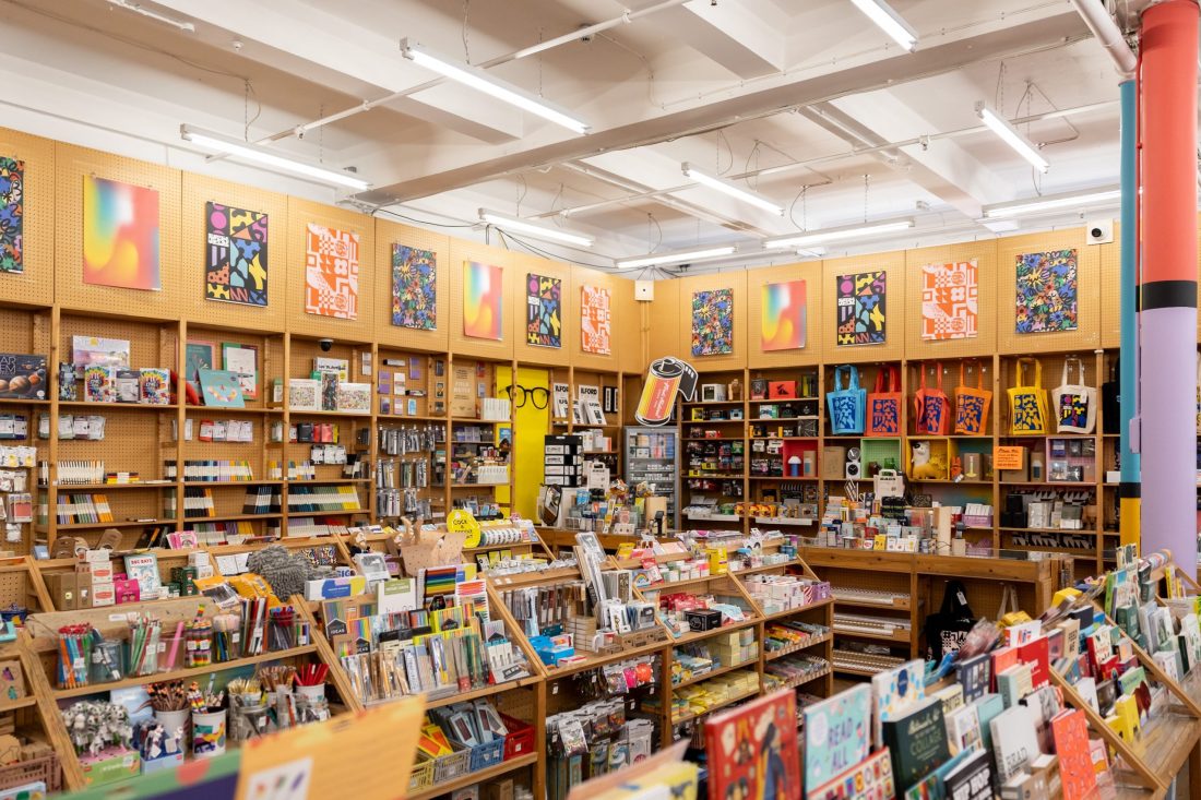 A brightly lit stationery and art supplies shop with shelves full of colourful supplies, books, and postcards. Vibrant abstract artwork is displayed on the walls above the shelves.