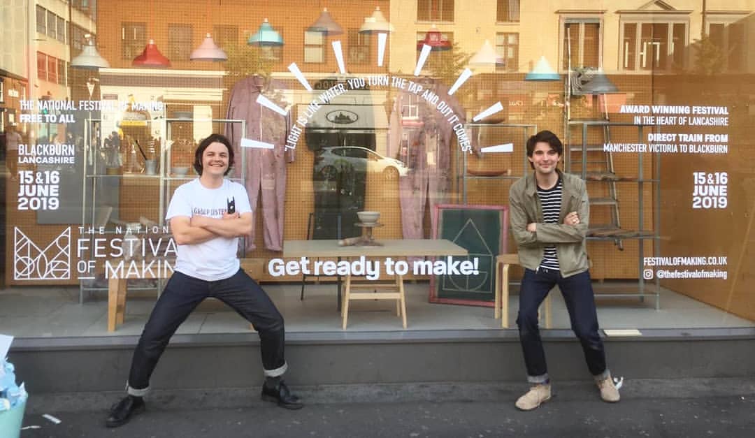 Two people pose and smile outside a large shop window decorated with signs for The National Festival of Making, taking place in Blackburn, Lancashire on 15–16 June 2019. The window reads, Get ready to make!.