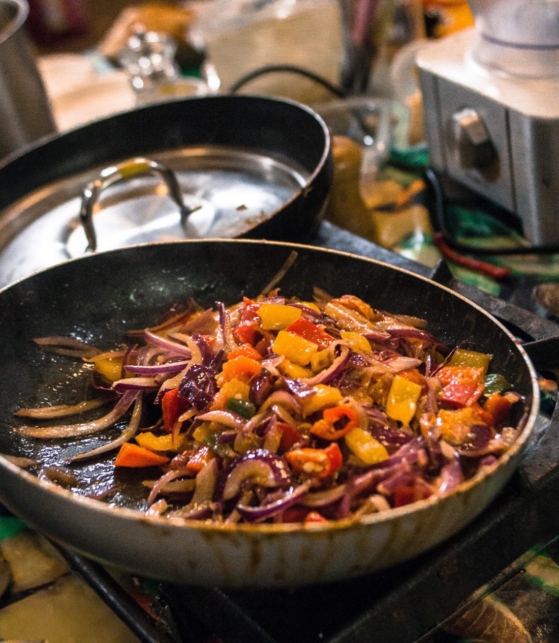 A black frying pan on a hob contains sautéed vegetables, including sliced red onions, yellow and red peppers, with a saucepan and kitchen appliances visible in the blurred background.