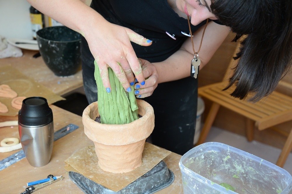 A person shapes green clay to resemble a cactus in a terracotta pot. Art supplies and containers are on the table, and the person has blue-painted nails and wears a key necklace.
