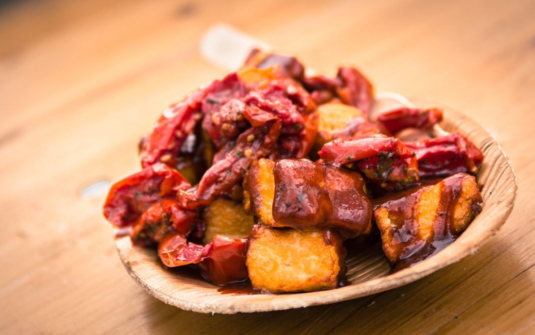 A wooden plate filled with crispy fried potato cubes topped with a spicy red sauce and chunks of red peppers, placed on a wooden table.
