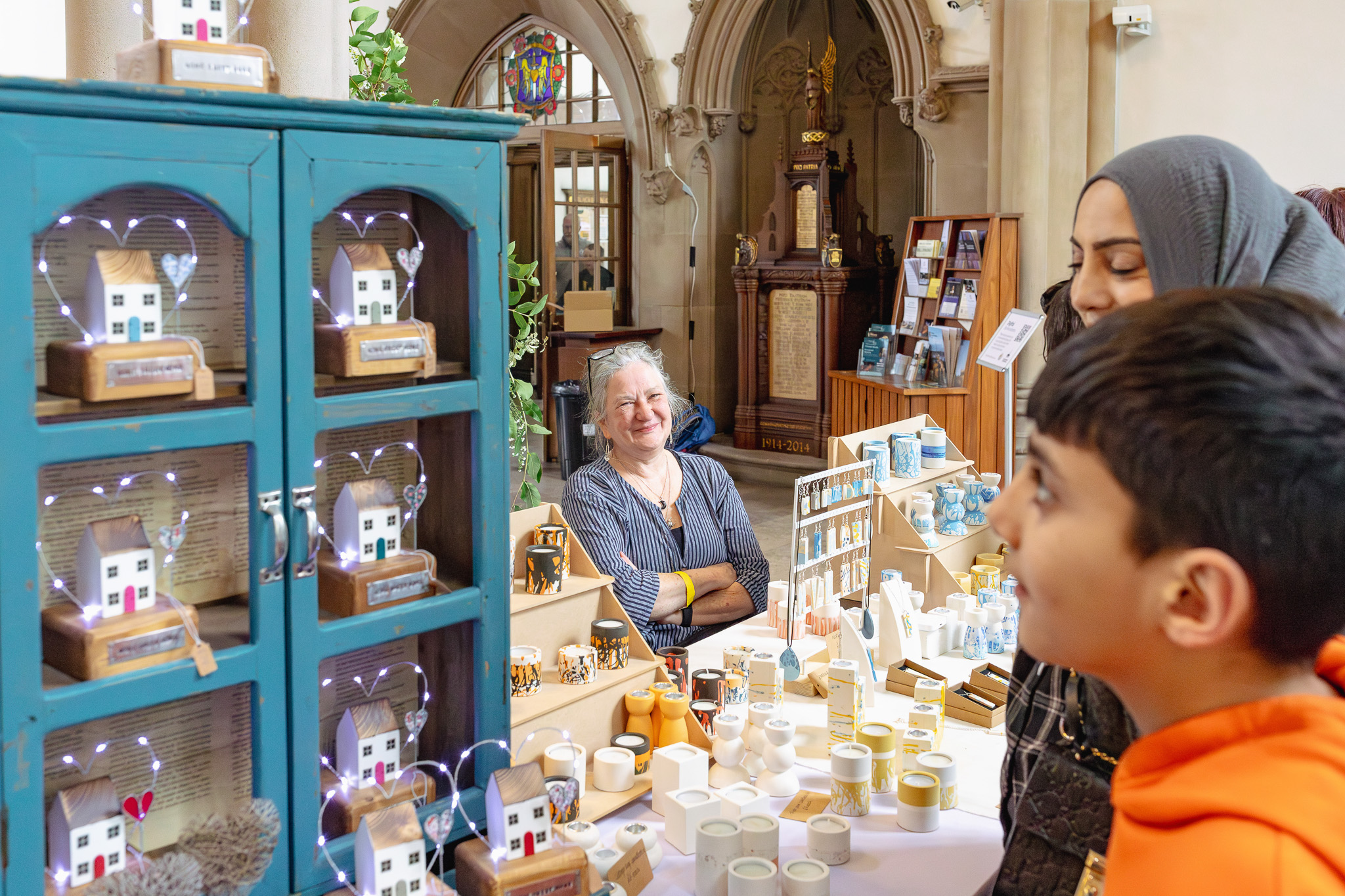 A market stall in a historic building displays handmade crafts. A woman smiles behind the table, while two visitors observe the art pieces with interest.