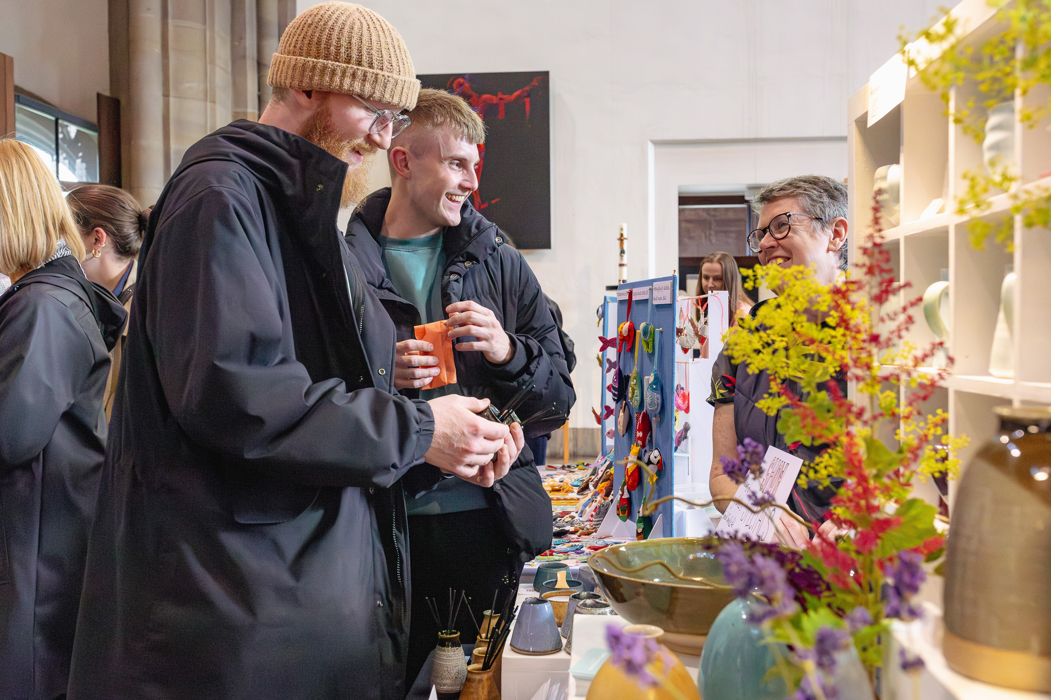 Two men in warm clothing admire crafts at a market stall. The vendor, smiling, engages with them.