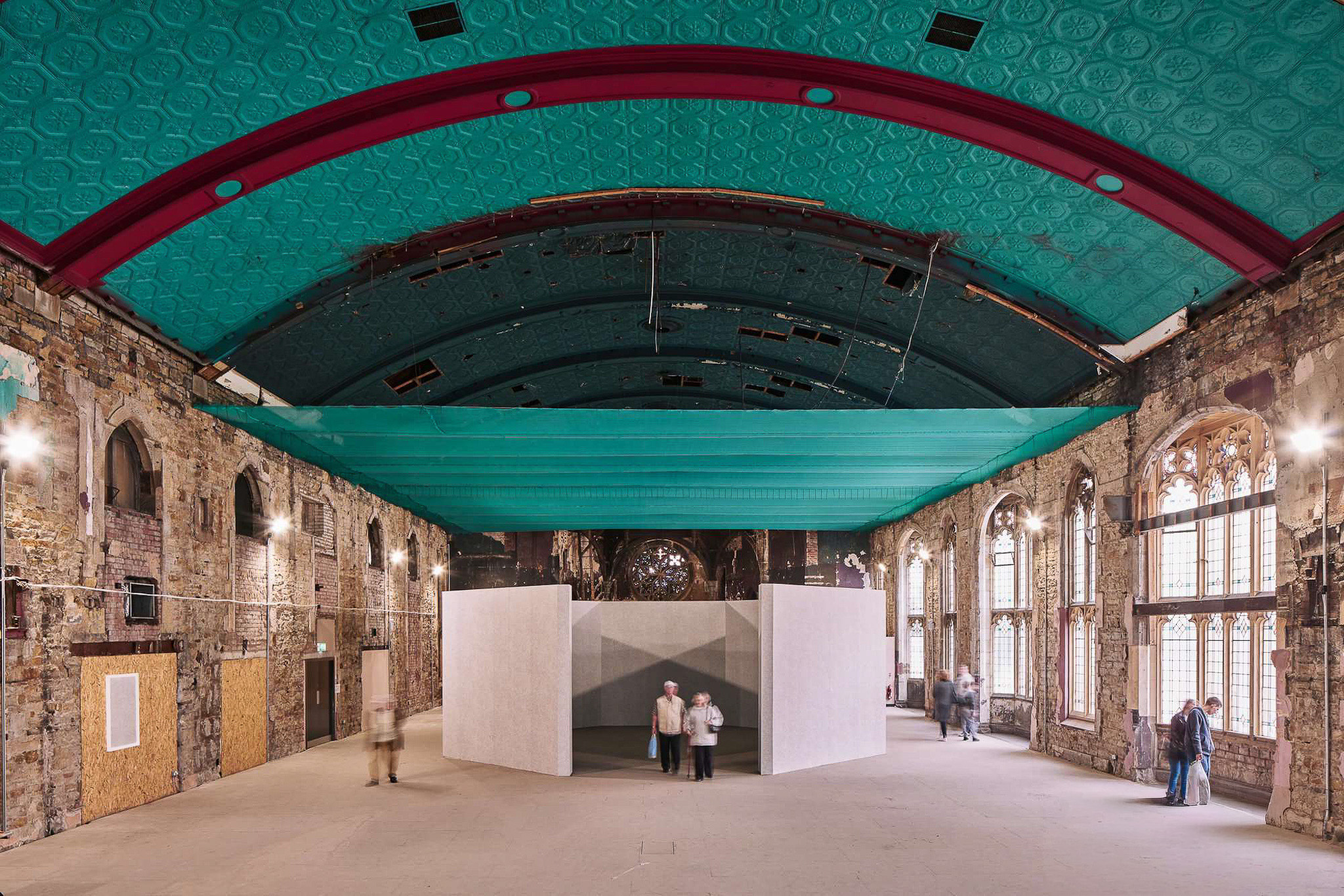 People walk inside a large, historic hall with exposed stone walls, arched windows, and a teal arched ceiling. A modern, white geometric structure stands at the centre of the space.