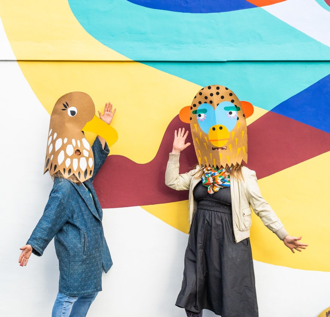 Two people wearing colourful animal masks and playful outfits pose joyfully in front of a bright, abstract mural with yellow, blue, and orange shapes. One mask is a bird; the other resembles a monkey.