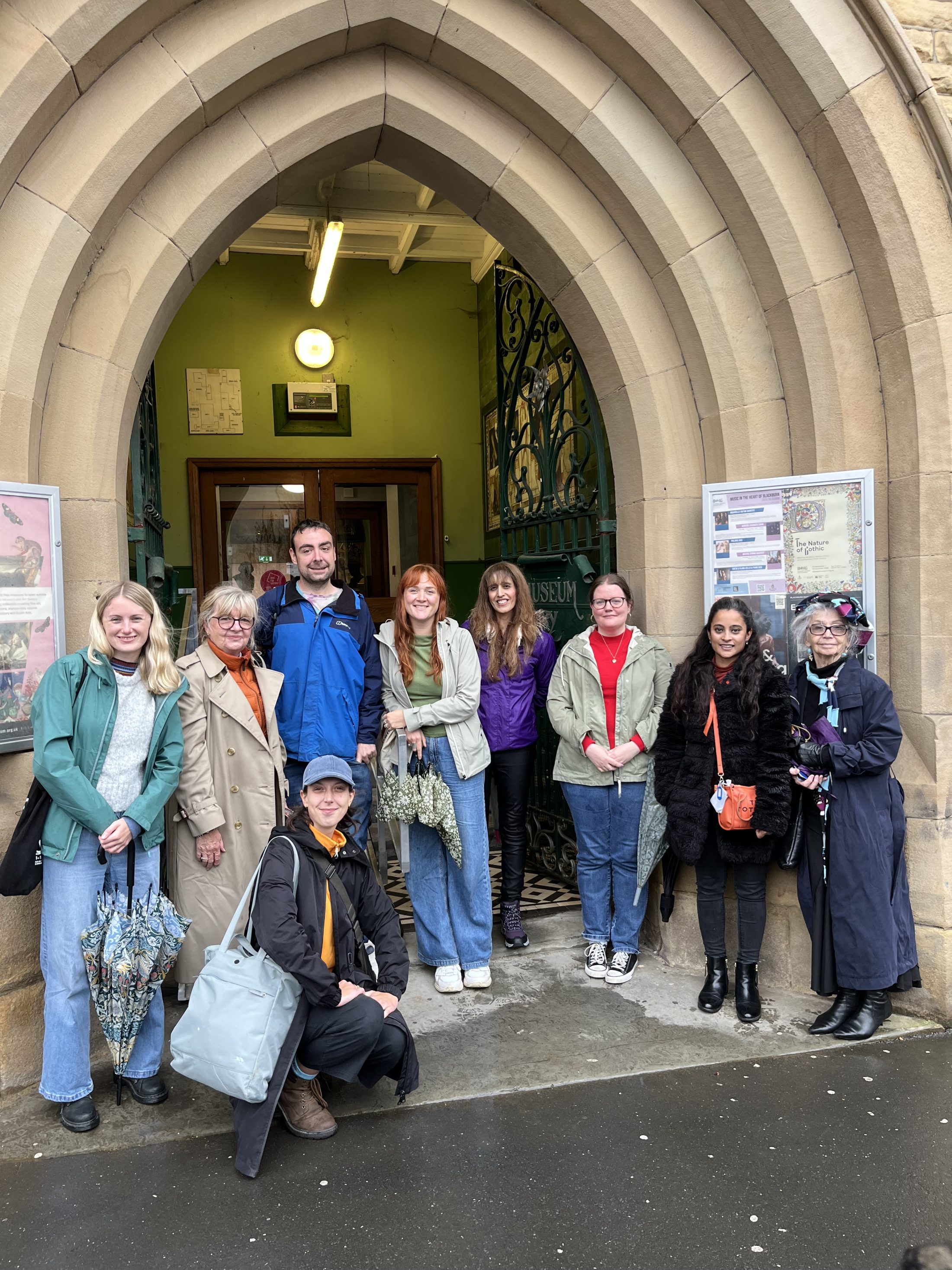 A group of nine people stand and kneel under a large stone archway at the entrance of a building, some smiling and dressed in coats. It appears to be a cloudy day, and the doorway leads into a museum.