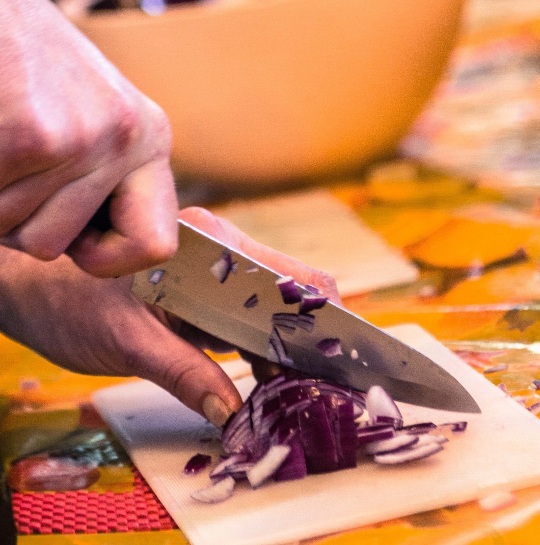 A person’s hands chopping a red onion with a large knife on a small chopping board. There is a blurred bowl in the background on a colourful tablecloth.