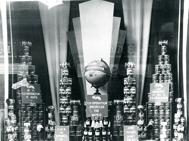 A black and white photo of a store window display of stacked cans and a central table top globe. Various signs promoting 'Co-Operation'.