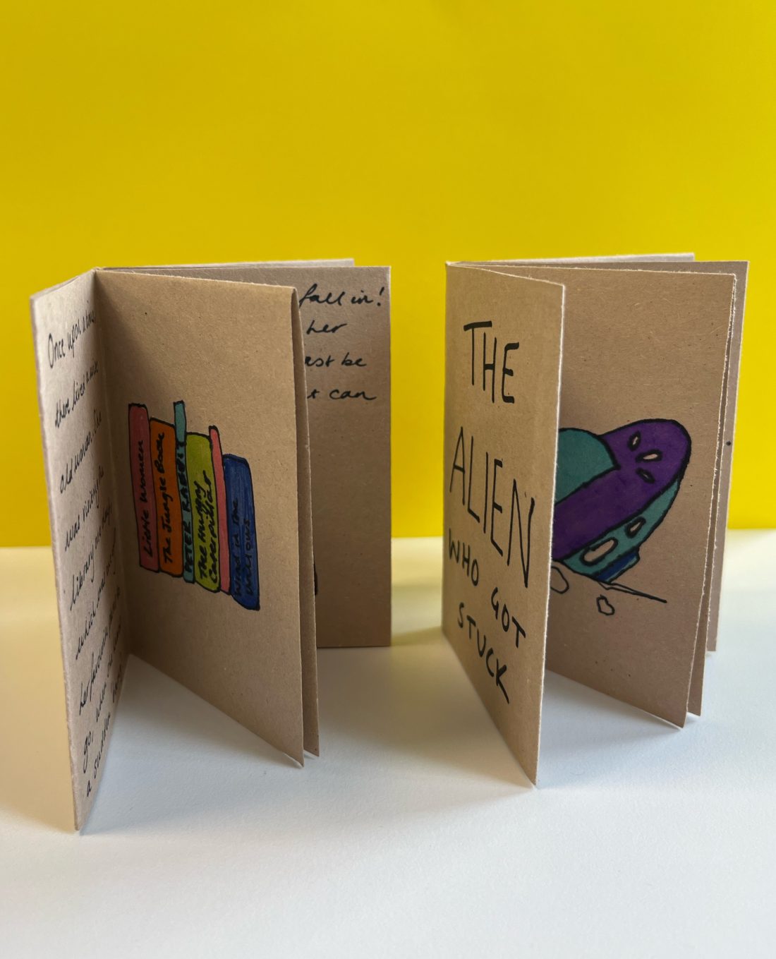 Two handmade books stand on a white surface against a yellow background. One cover reads "The Alien Who Got Stuck" with a drawing of a spaceship. Pages contain sketches and text.