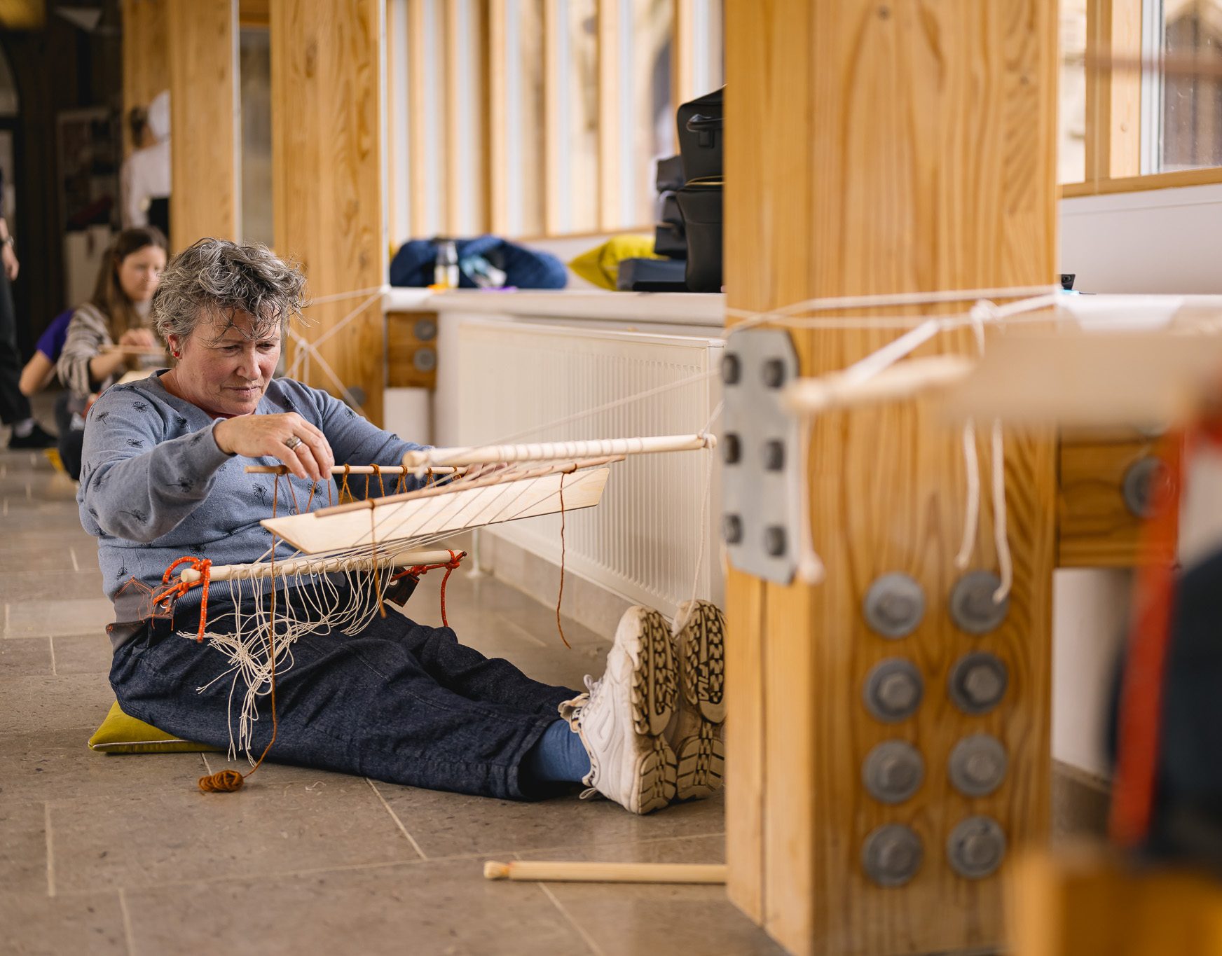 A person sits on the floor, weaving at a handmade loom attached to wooden beams. They are focused on their work, with string and weaving tools around them.