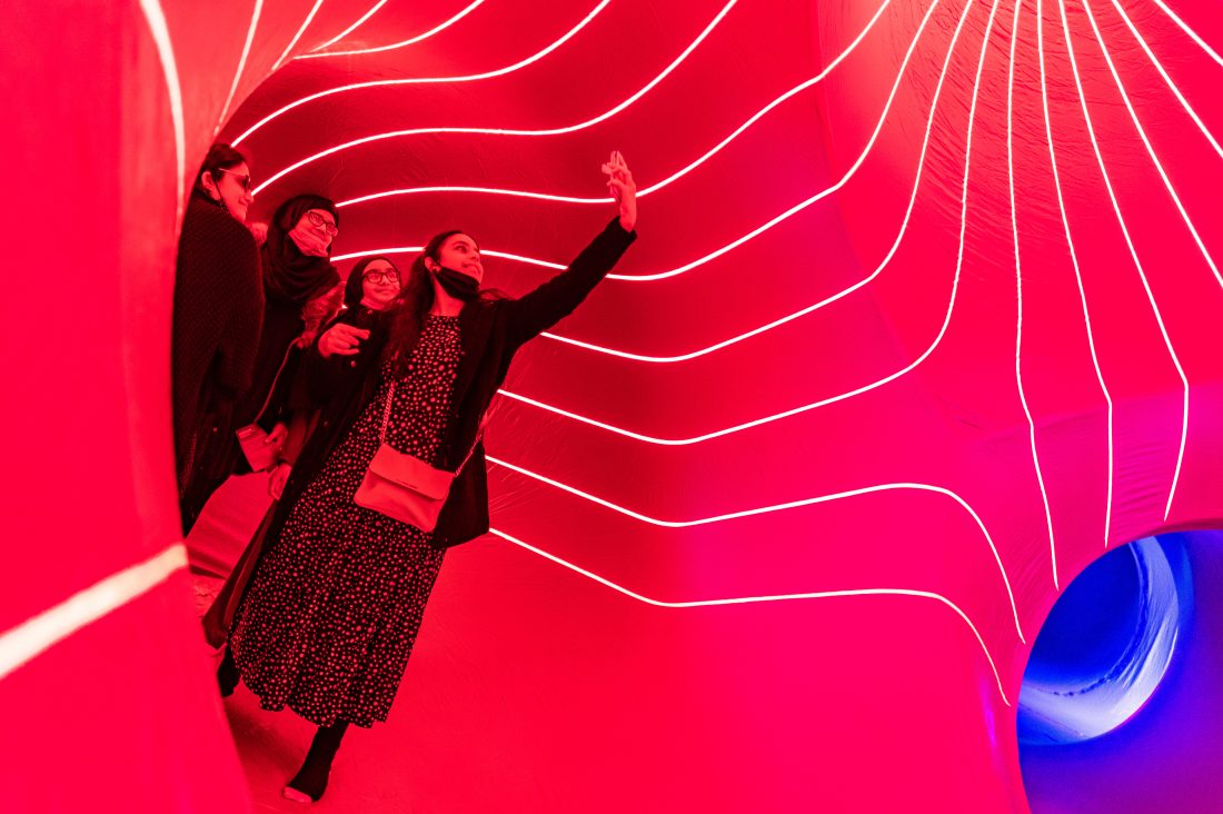 Four people walk through a tunnel-like installation illuminated with bright red lights and curved glowing lines; one person in front takes a selfie, while the others follow and look around curiously.