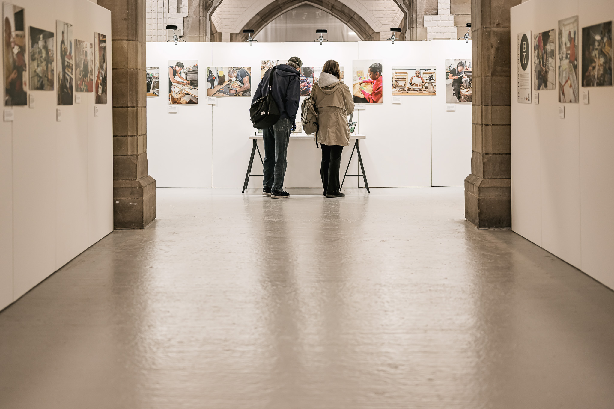 Two people stand closely together, viewing artwork displayed on a white wall in a spacious, well-lit gallery with high arches and photographs hanging on both side walls.