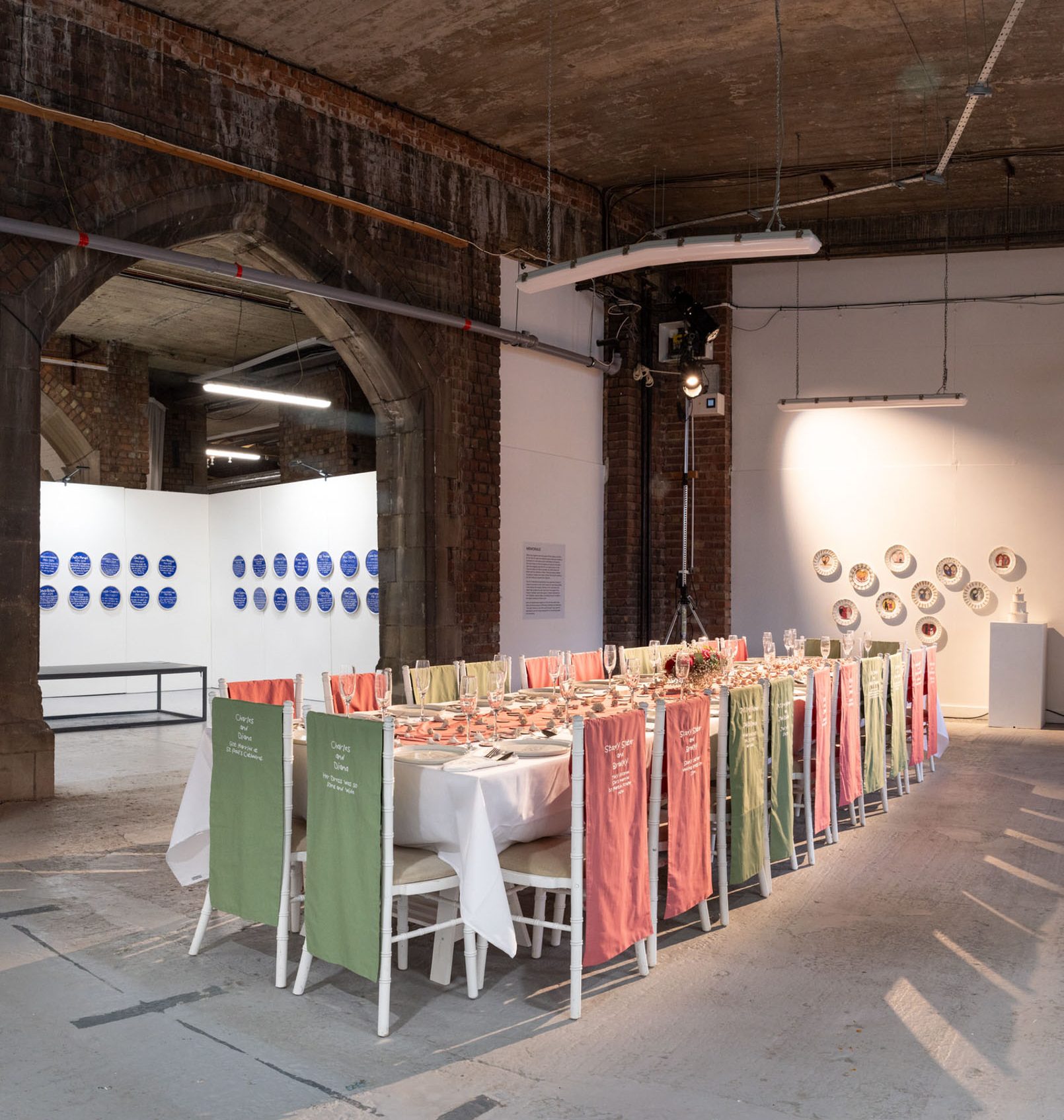 A long dining table with pink and green fabric draped over white chairs is set for a meal in an industrial-style gallery space, with plates displayed on white walls in the background.