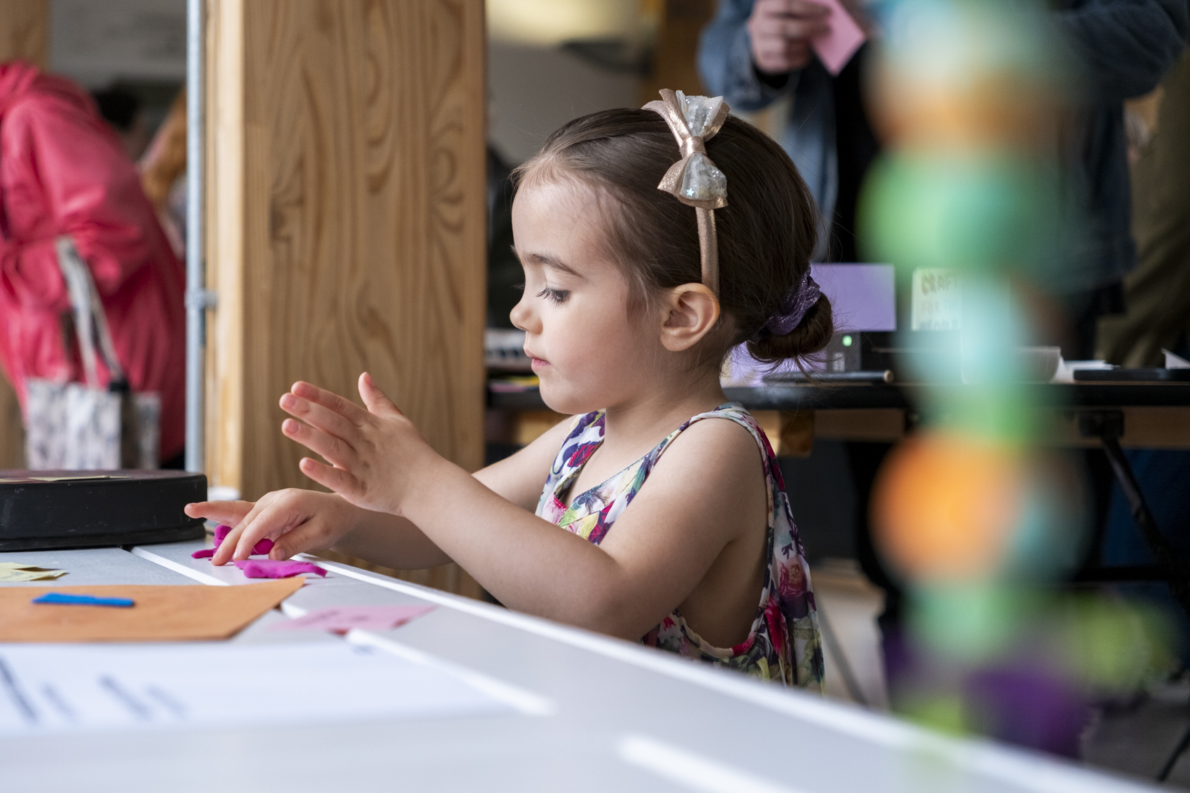 Young girl in floral dress and bow headband crafts at a table.