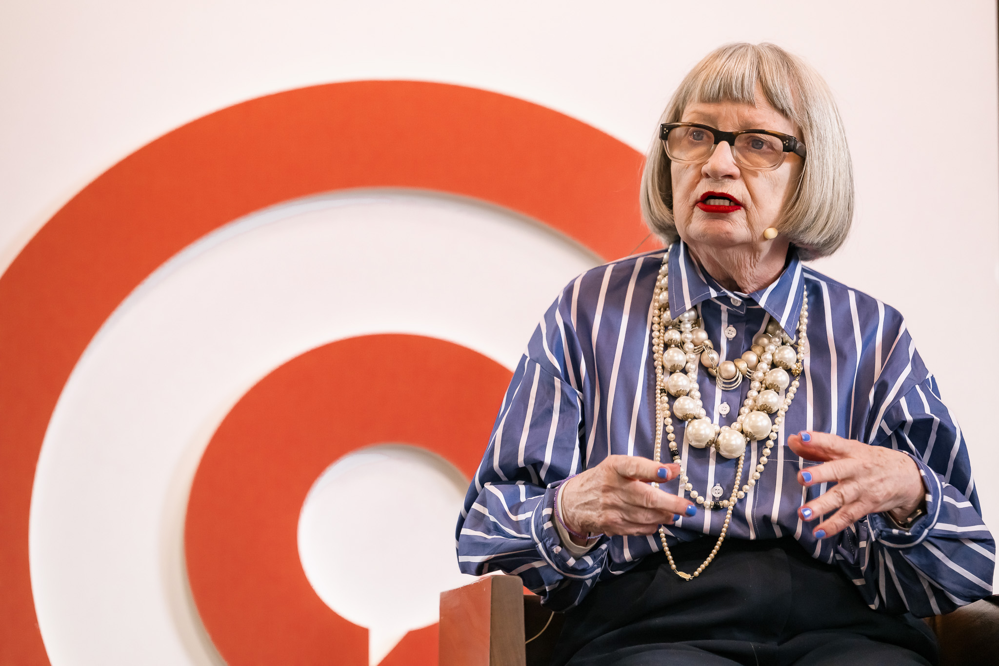 Esme Young with grey bobbed hair, red lipstick, and glasses speaks while seated. She wears a blue striped shirt, large beaded necklaces, and a headset microphone. A red spiral design is visible in the background.