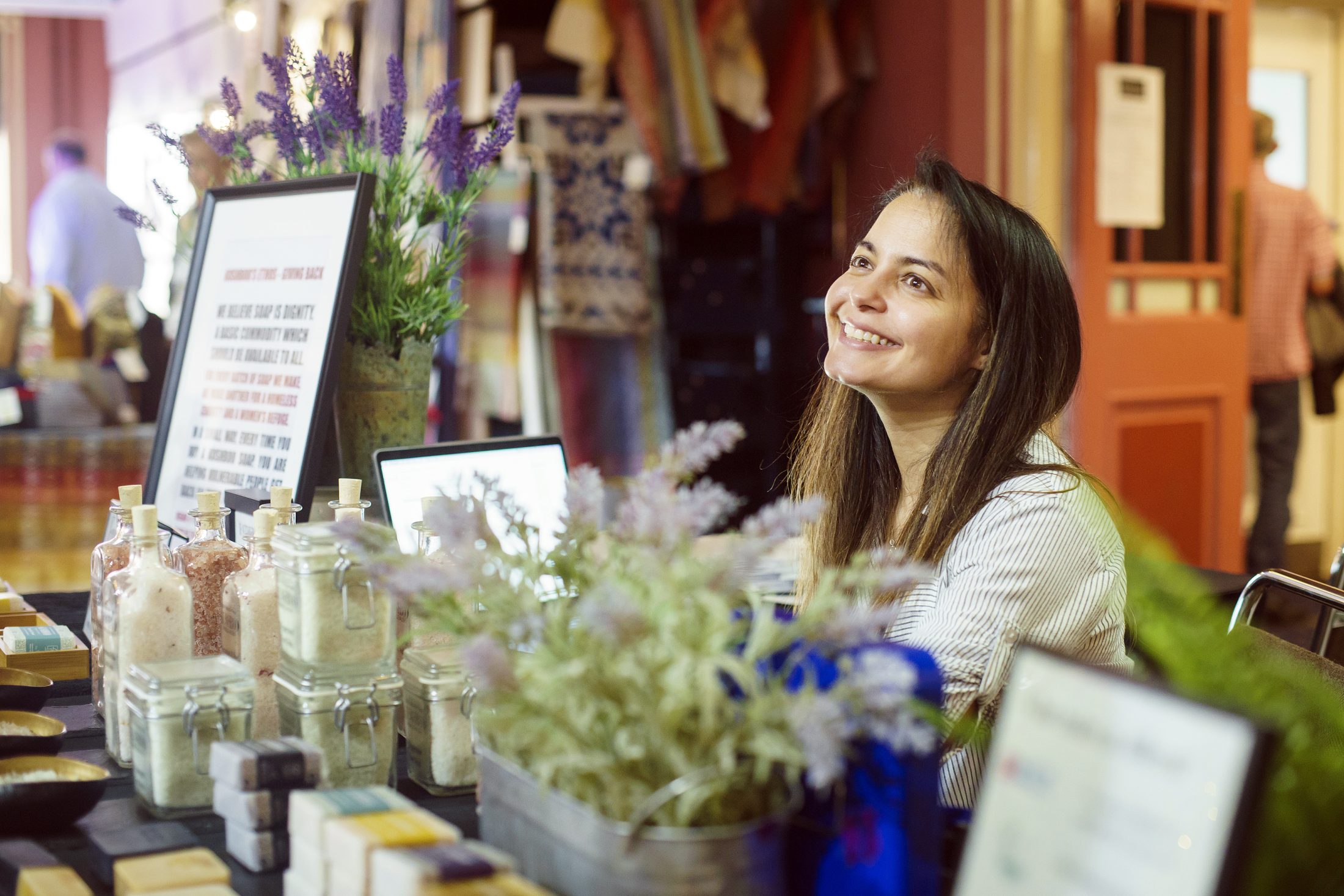 Smiling woman at a market stall with homemade soap and jars, surrounded by flowers and a sign.