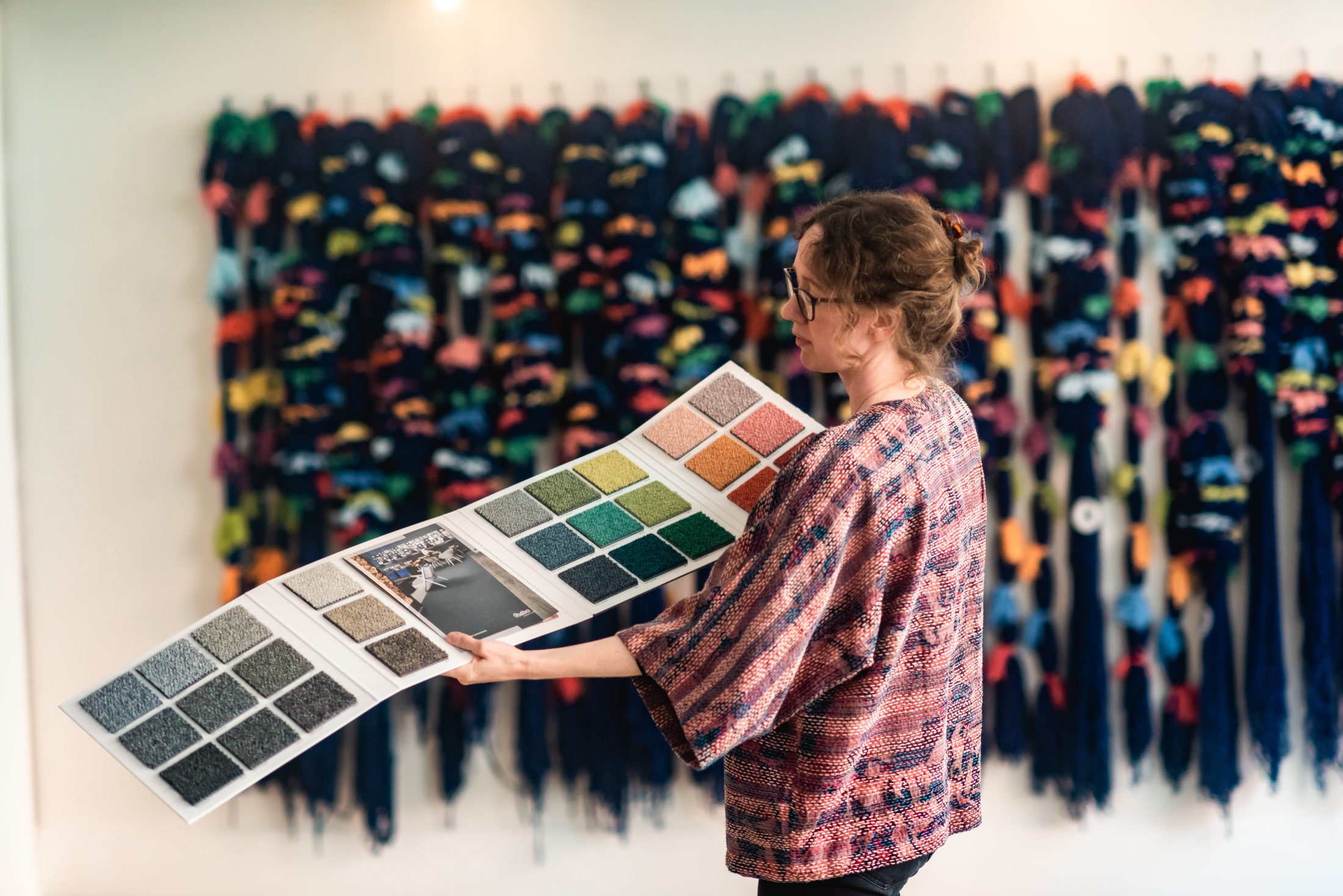 Woman in a patterned shirt holds a colour swatch book, examining vibrant textile samples, with colourful hanging artwork in the background.