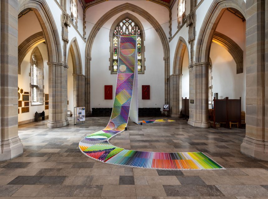 A large, colourful textile installation made of hundreds of fabric tubes arranged in a winding path stretches from the floor up to a tall window inside a grand church with high vaulted ceilings and stone arches.