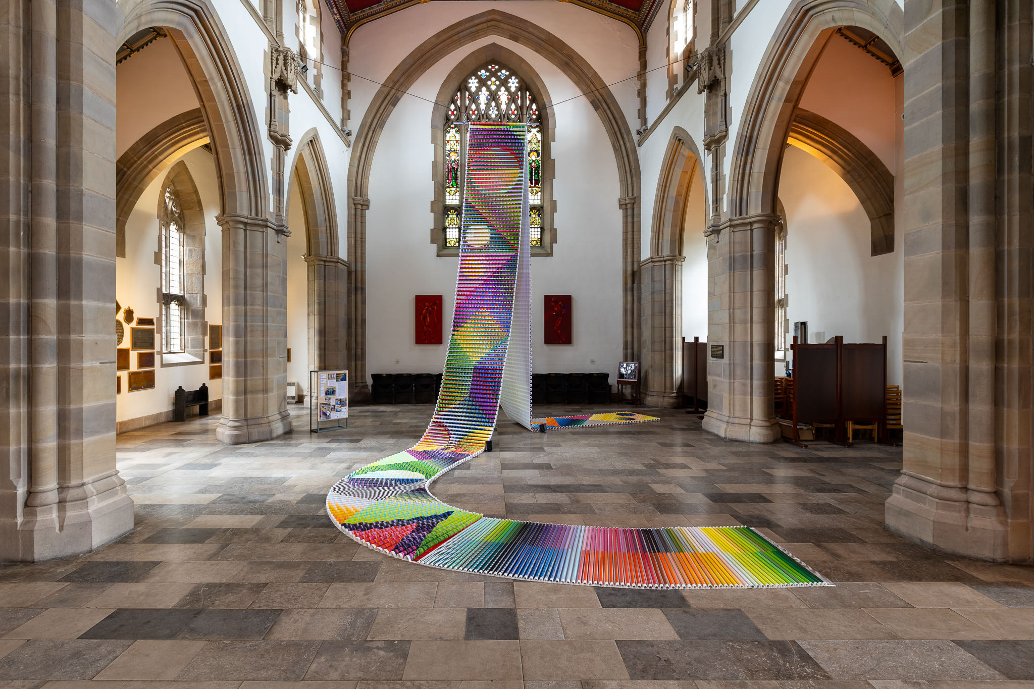 A large, colourful textile installation made of hundreds of fabric tubes arranged in a winding path stretches from the floor up to a tall window inside a grand church with high vaulted ceilings and stone arches.