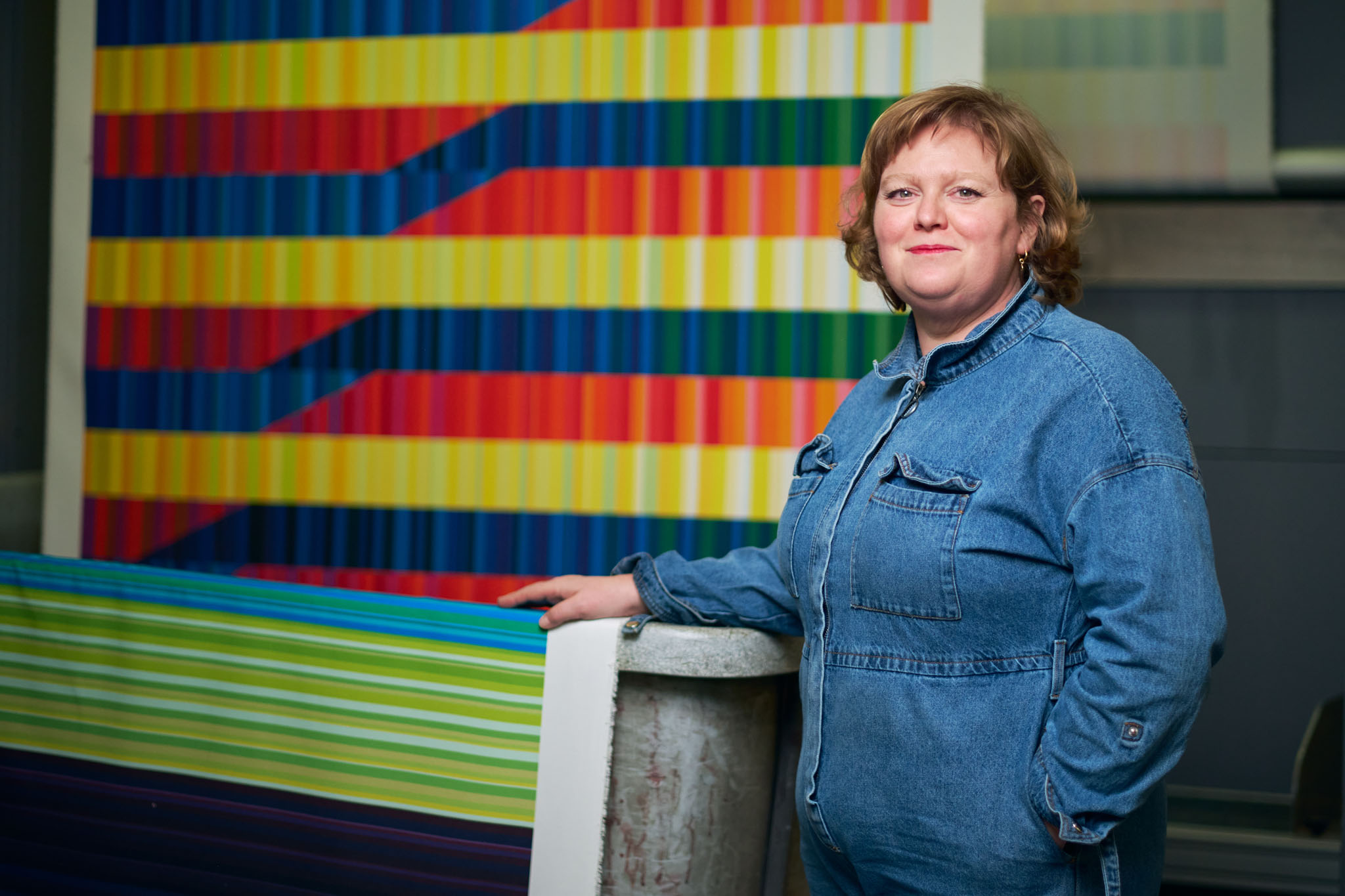 A woman in a denim jacket stands beside large, colourful abstract art featuring bold horizontal and diagonal stripes in various bright colours. She is smiling and looking at the camera.