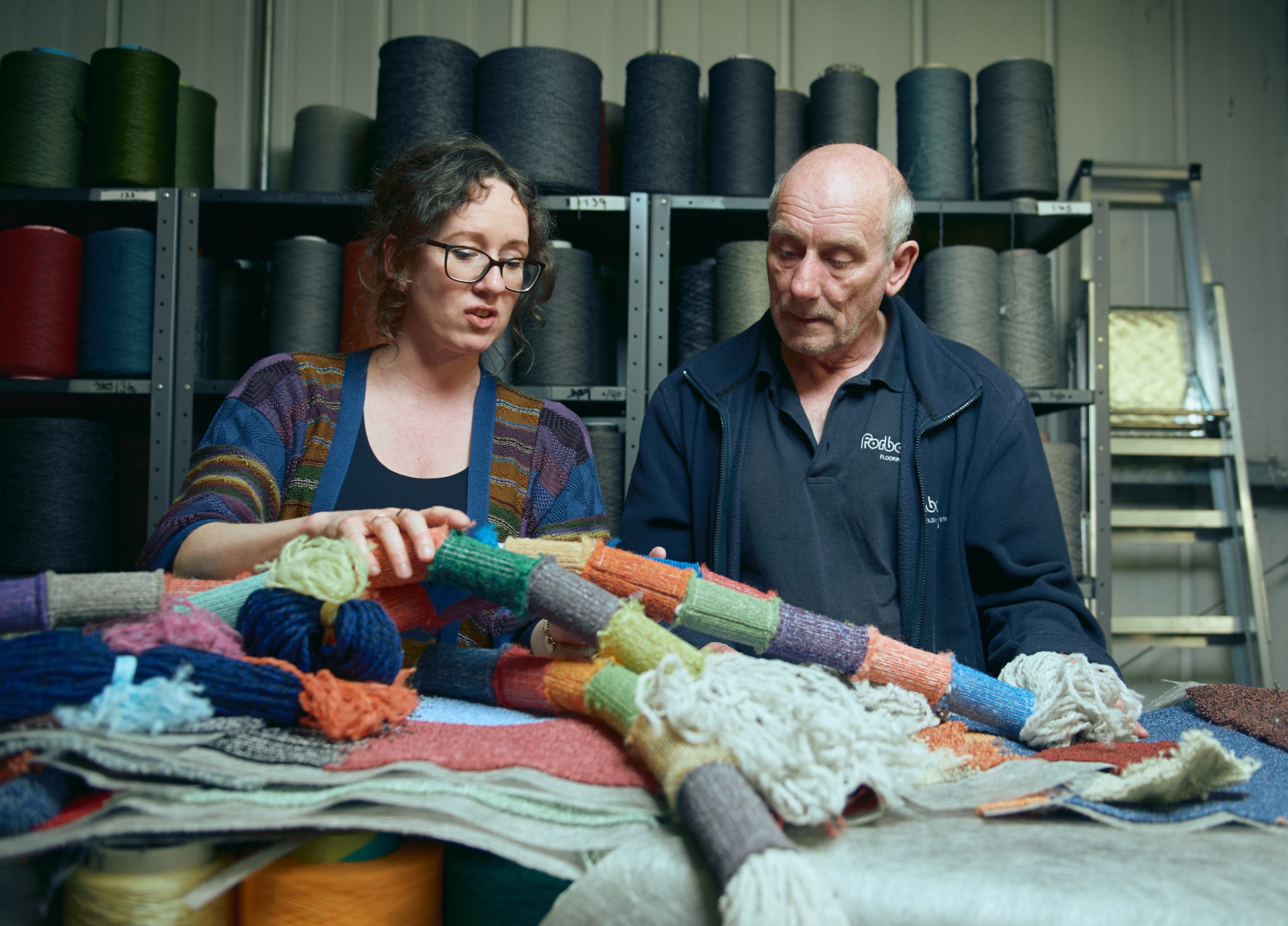 A woman and an older man sit at a table covered with colourful yarn and knitted fabric samples, discussing their work in a yarn-filled workshop with shelves and a ladder in the background.