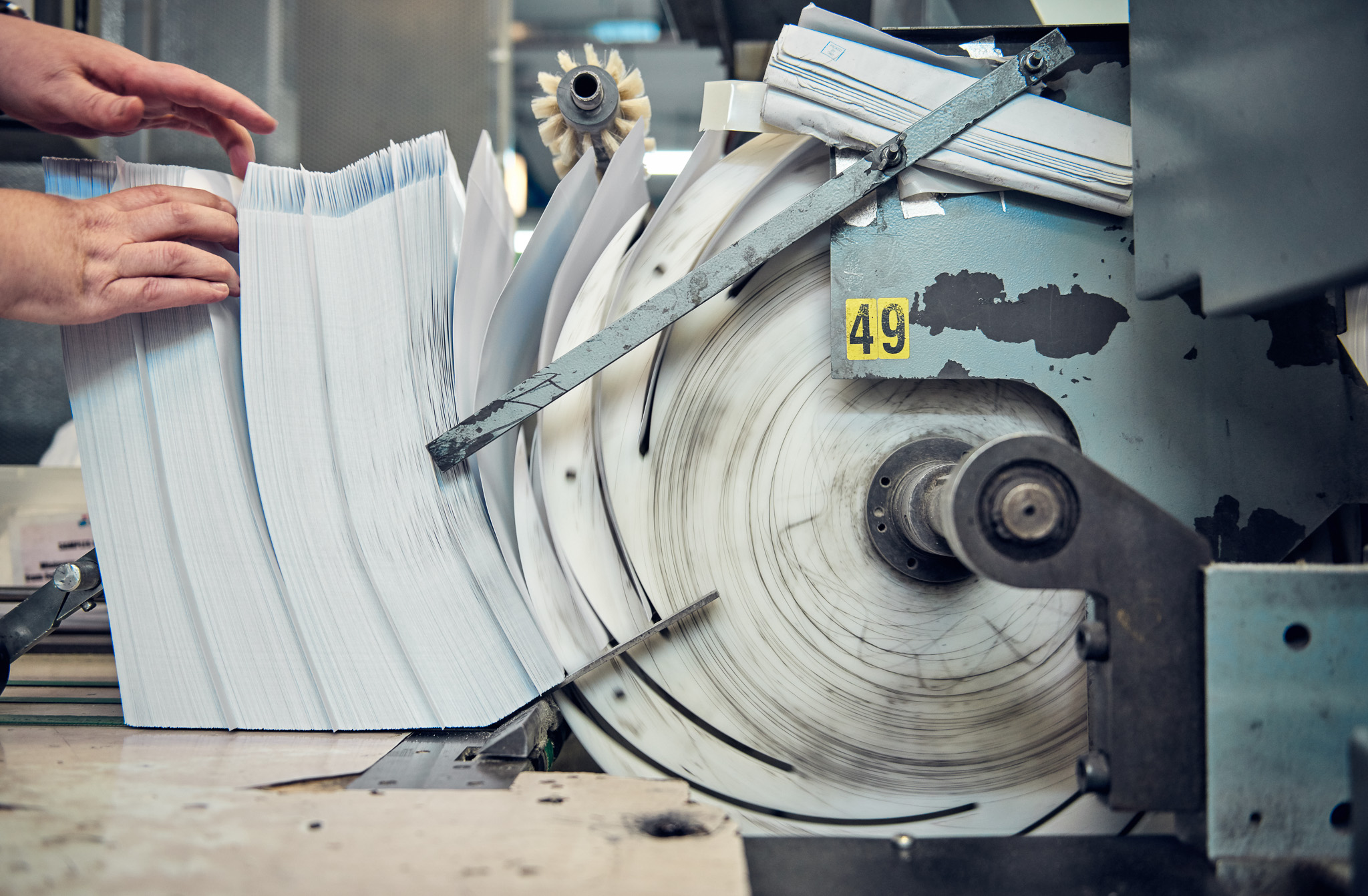 Hands hold stacks of paper beside a large, spinning printing press labeled with the number 49.