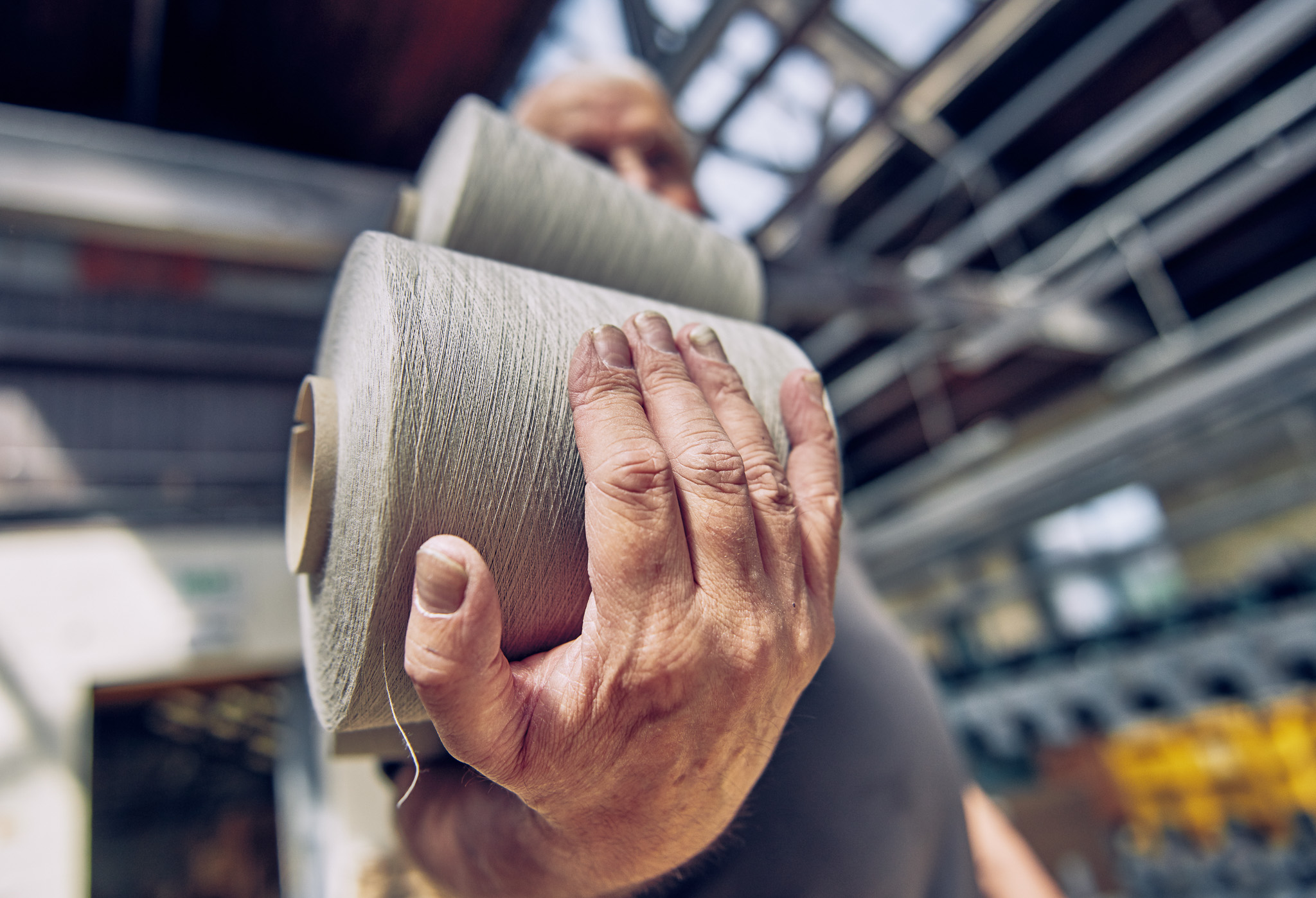 A person holds two large reels of thread in an industrial setting, with their hand and the reels in sharp focus and the background blurred in a textile manufacturing facility.
