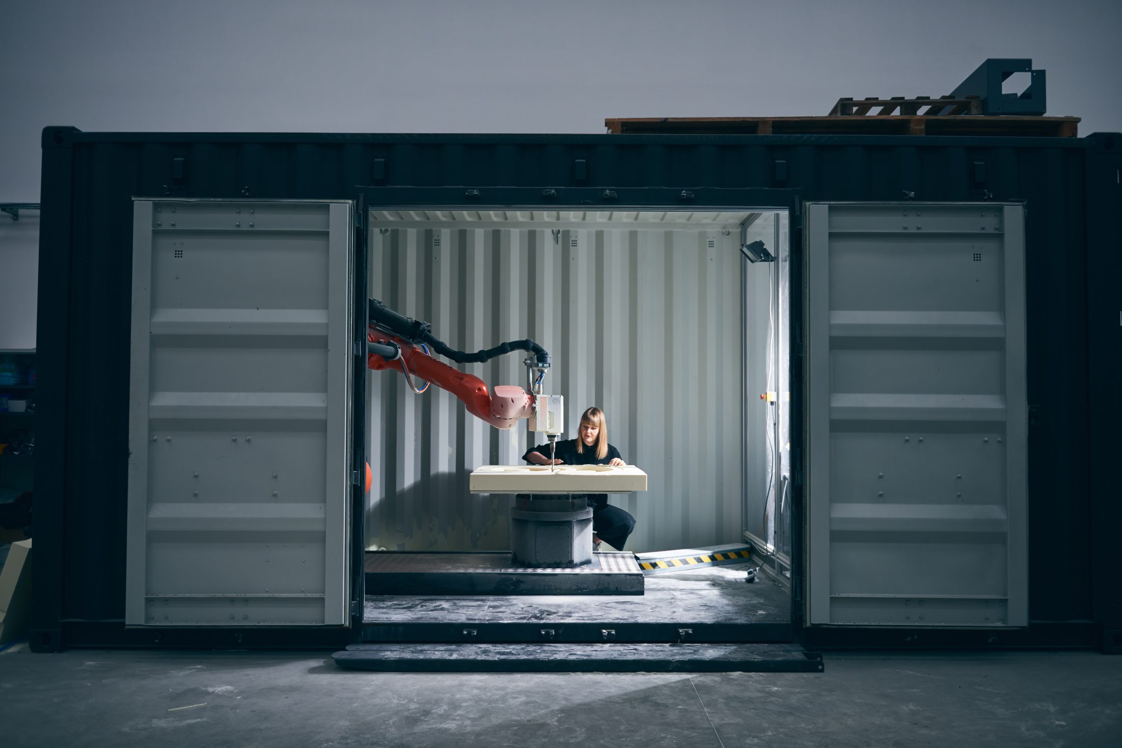 A person works on a project inside a shipping container, using a large industrial robotic arm. The container doors are open, and the setting appears to be an industrial or workshop space.