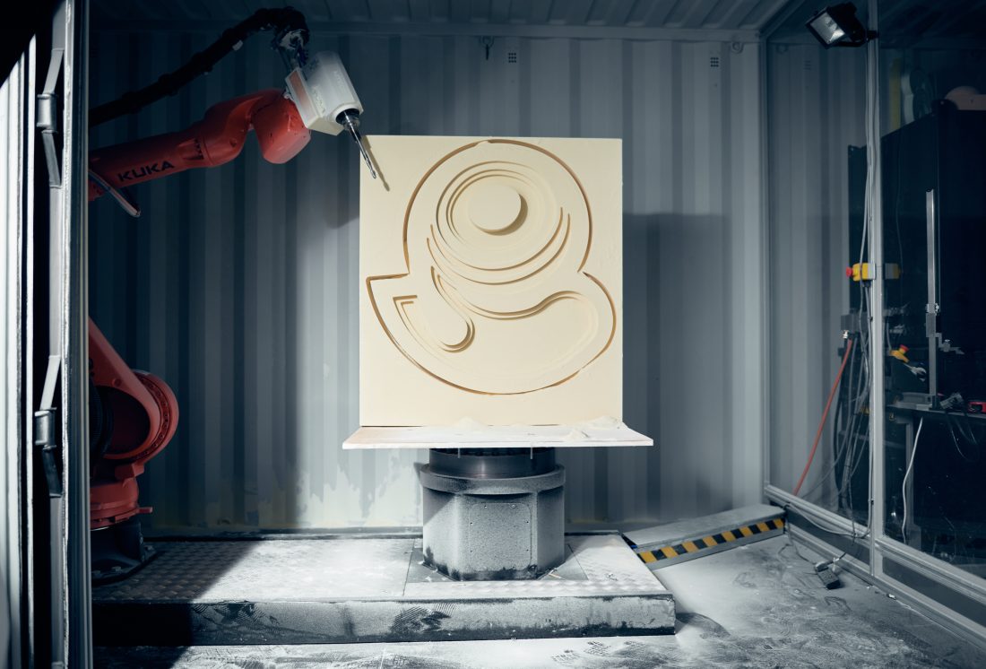 A robotic arm carves a swirling pattern into a large, square panel.