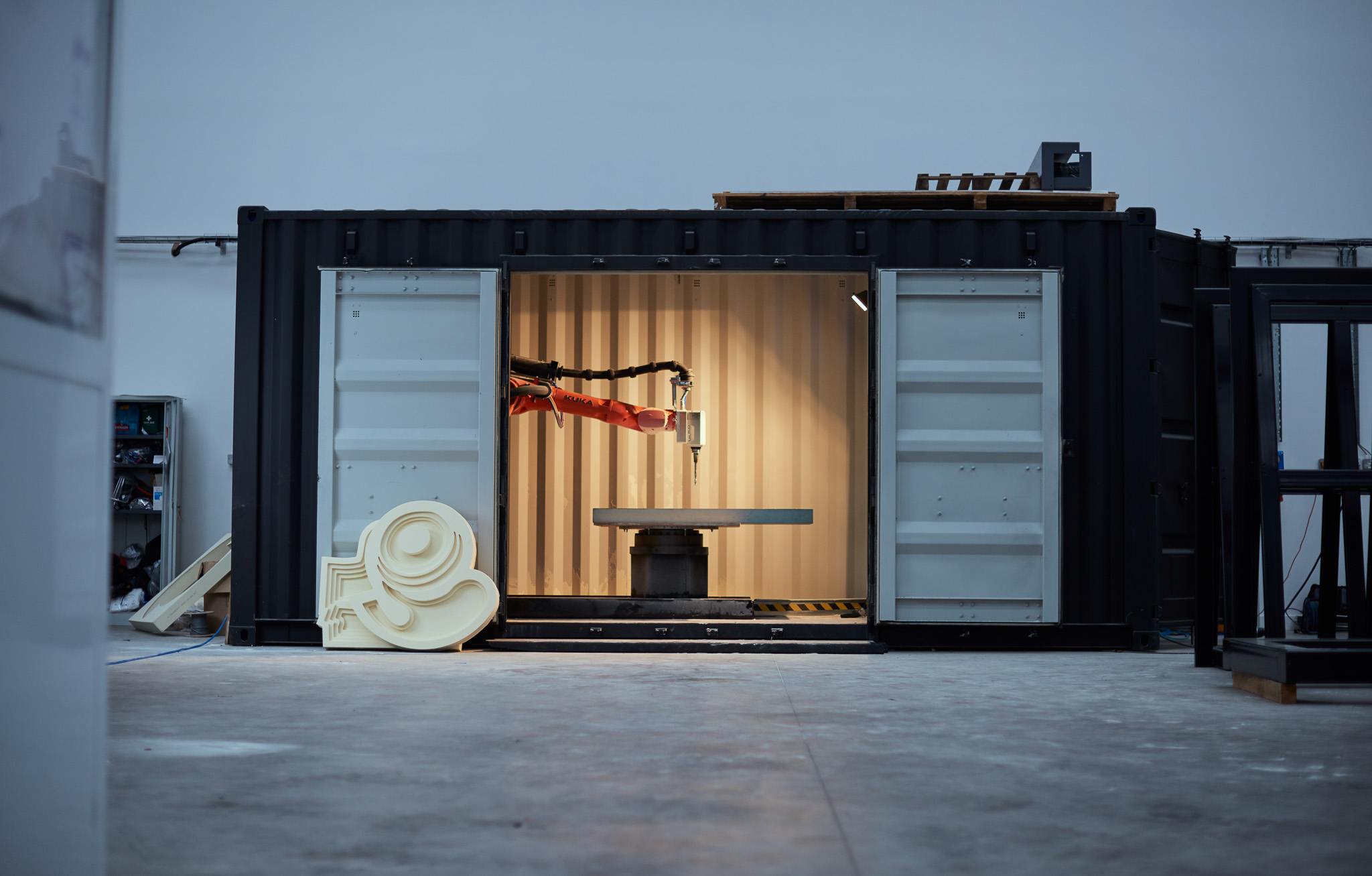 A robotic arm operates inside an open shipping container, positioned over a circular platform. Large 3D-printed artworks are placed on the floor nearby in a spacious industrial setting.