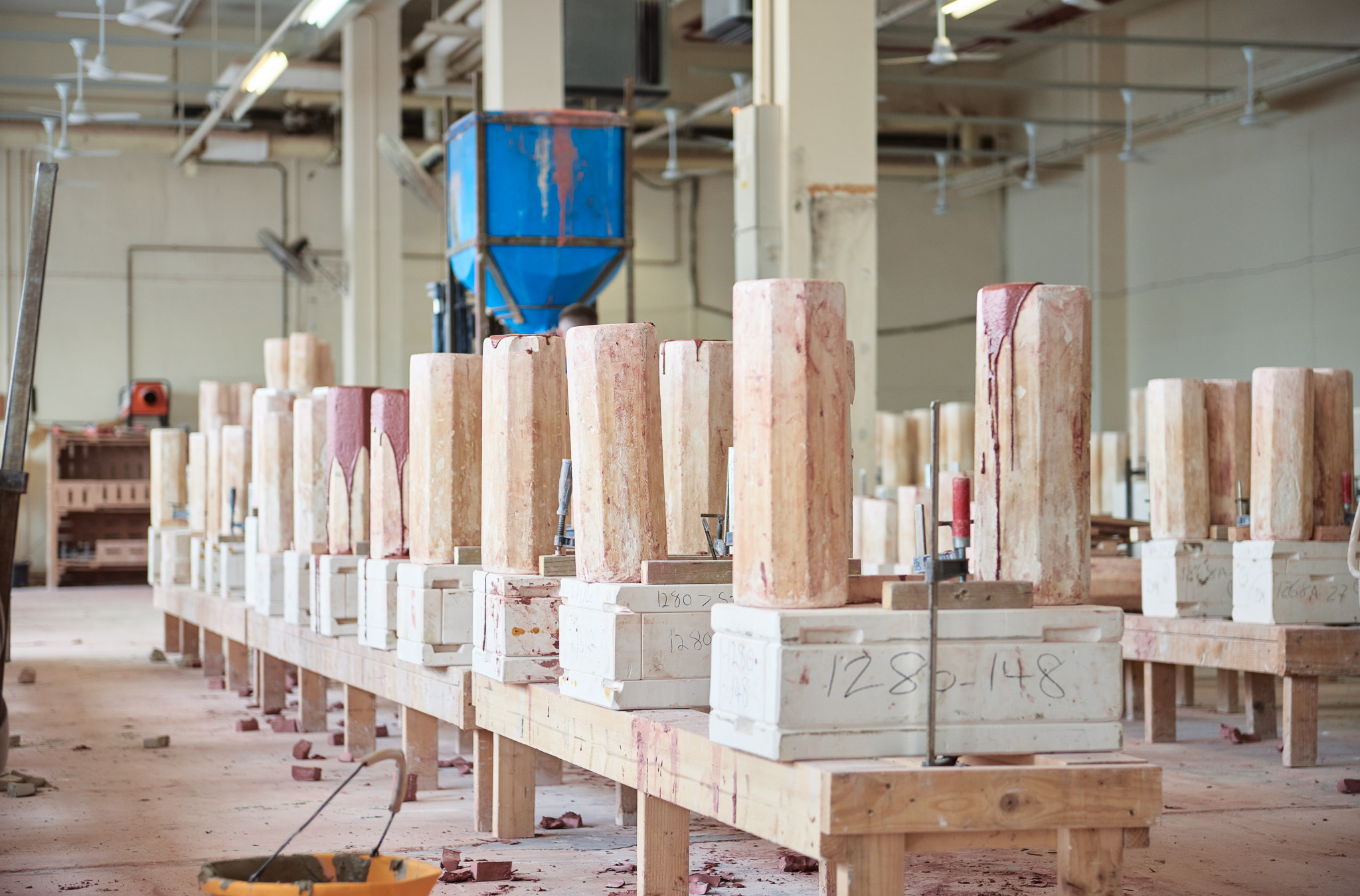 Rows of tall, round ceramic moulds with liquid clay flowing over the edges are secured onto workbenches in a large, bright workshop. Various machines and tools are in the background.