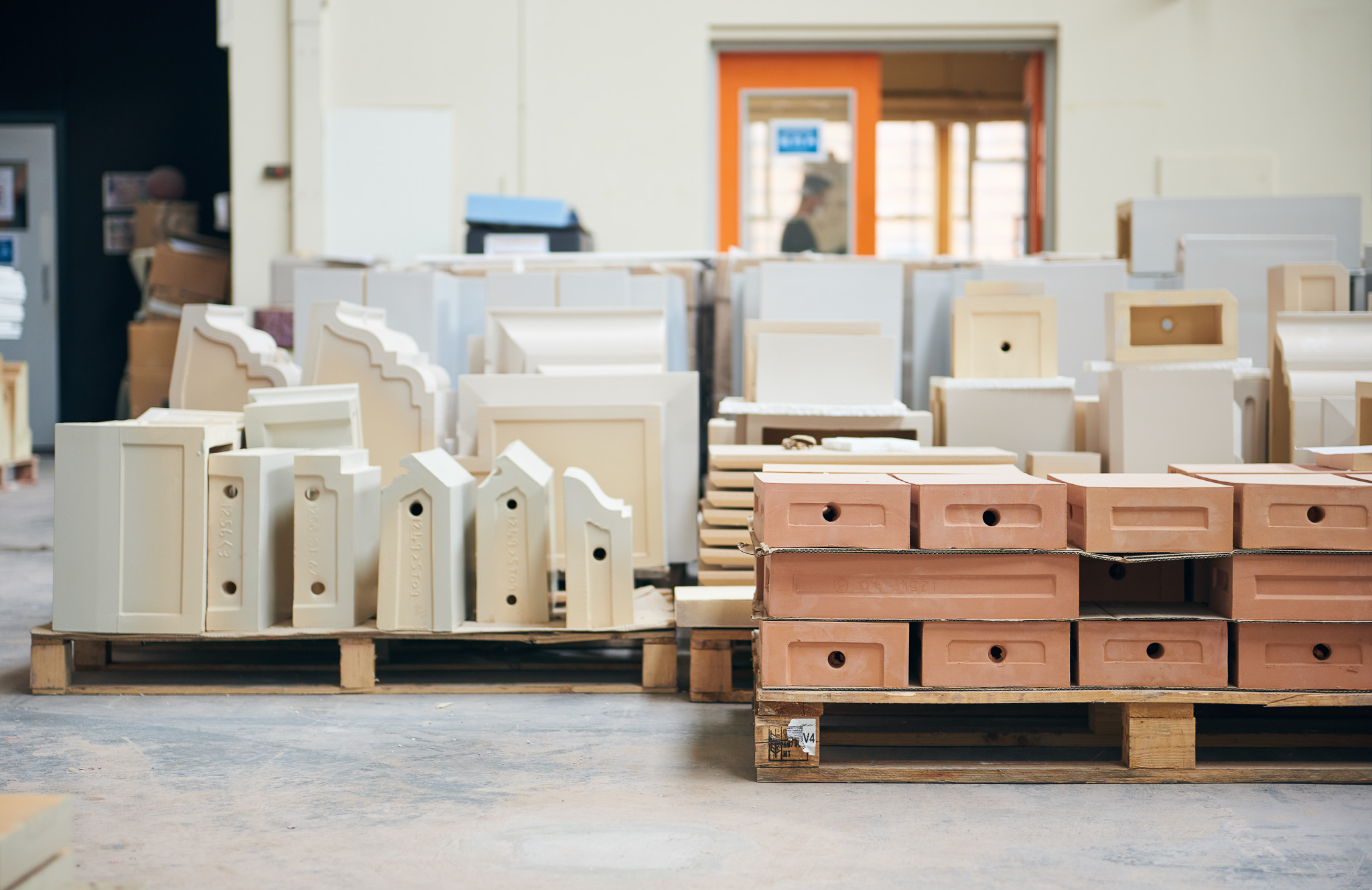 Stacks of ceramic moulds and pottery forms are organised on wooden pallets in a spacious workshop with a person visible in the background through an open orange-framed door.