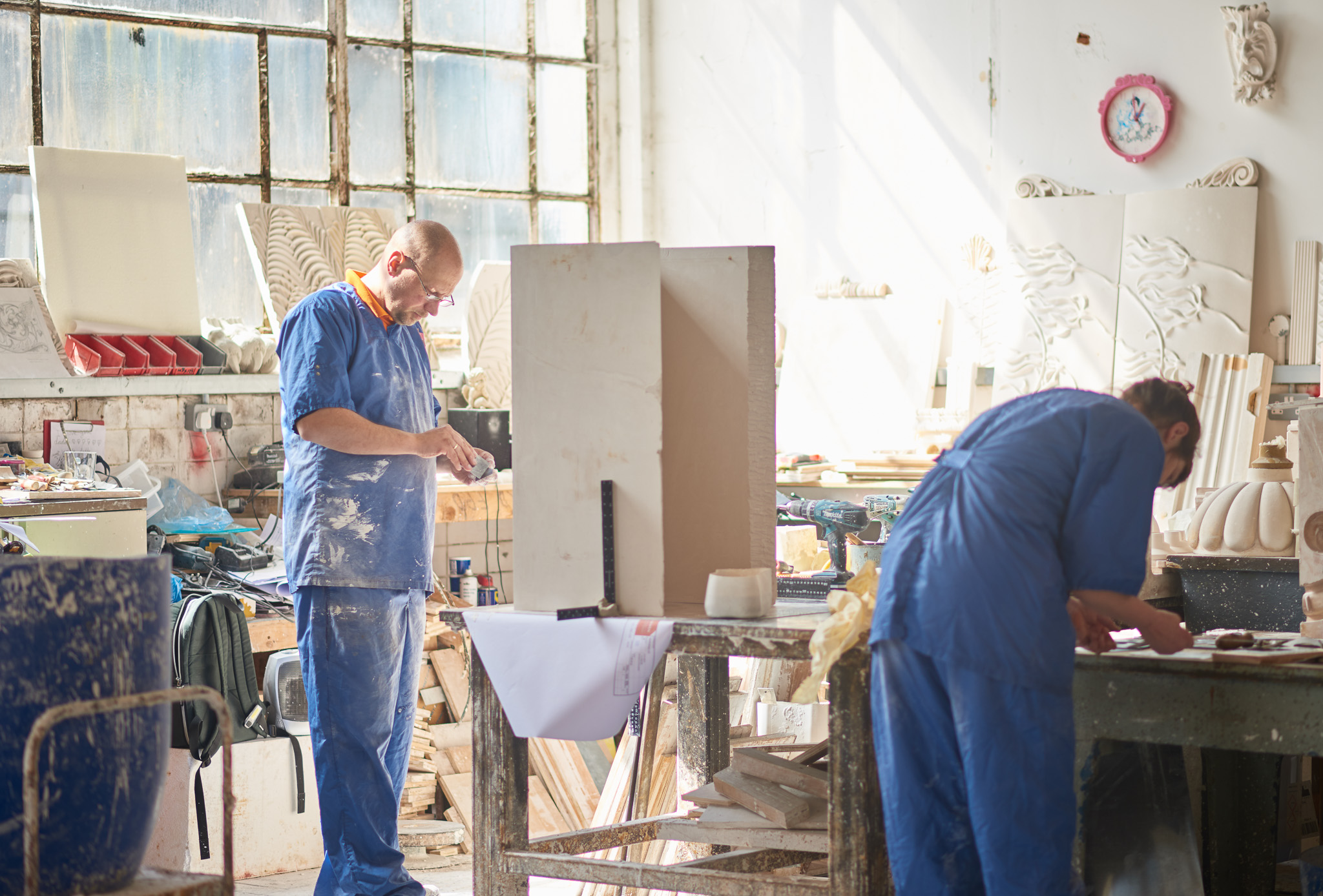 Two people in blue overalls work in a bright, sunlit studio filled with sculptures, tools, and supplies. Dust and materials are scattered on tables and the floor.