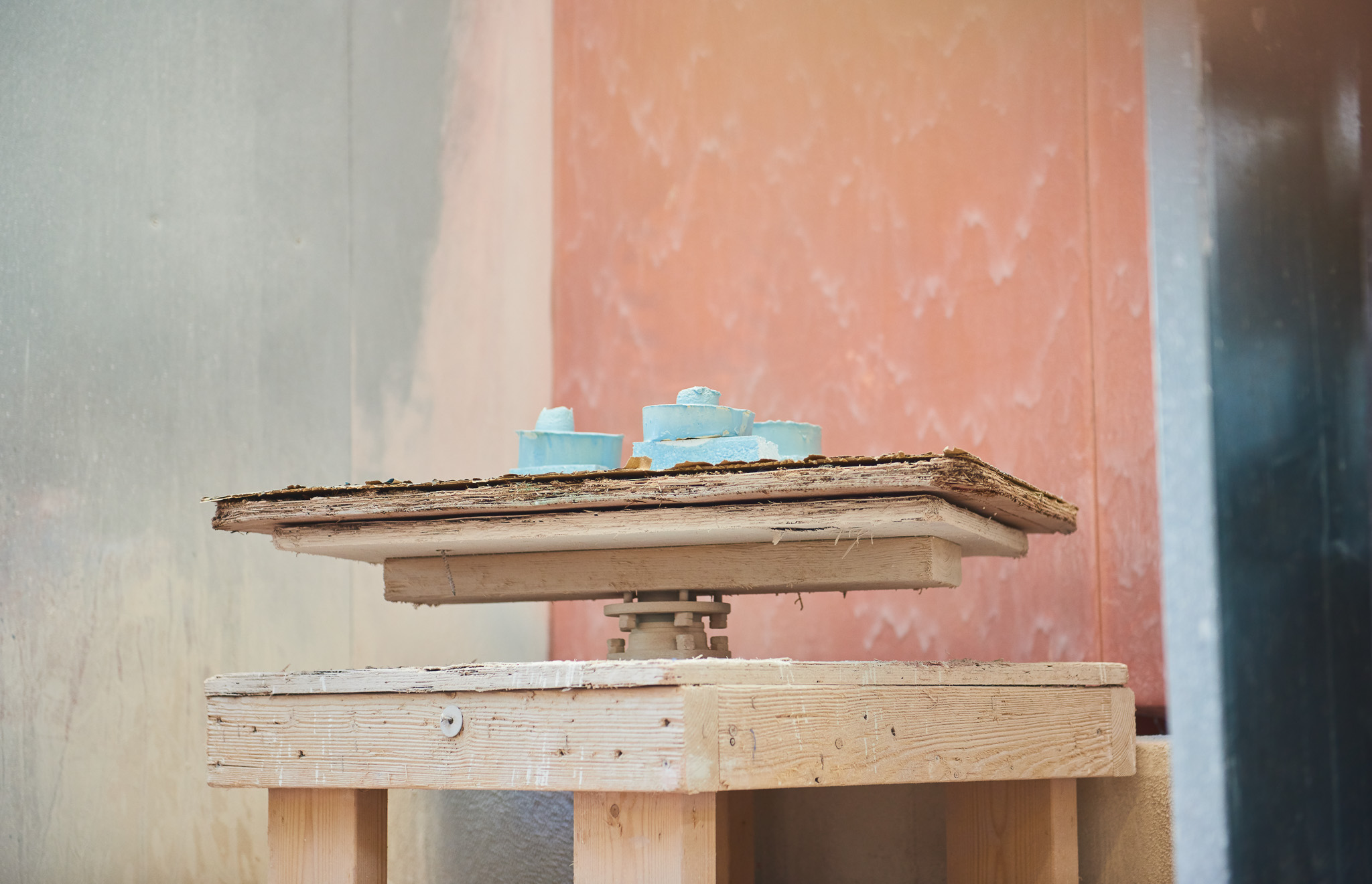 A small sculpture made of blue material sits on a textured brown platform, which is mounted on a wooden stand against a background of softly coloured walls in shades of peach and grey.