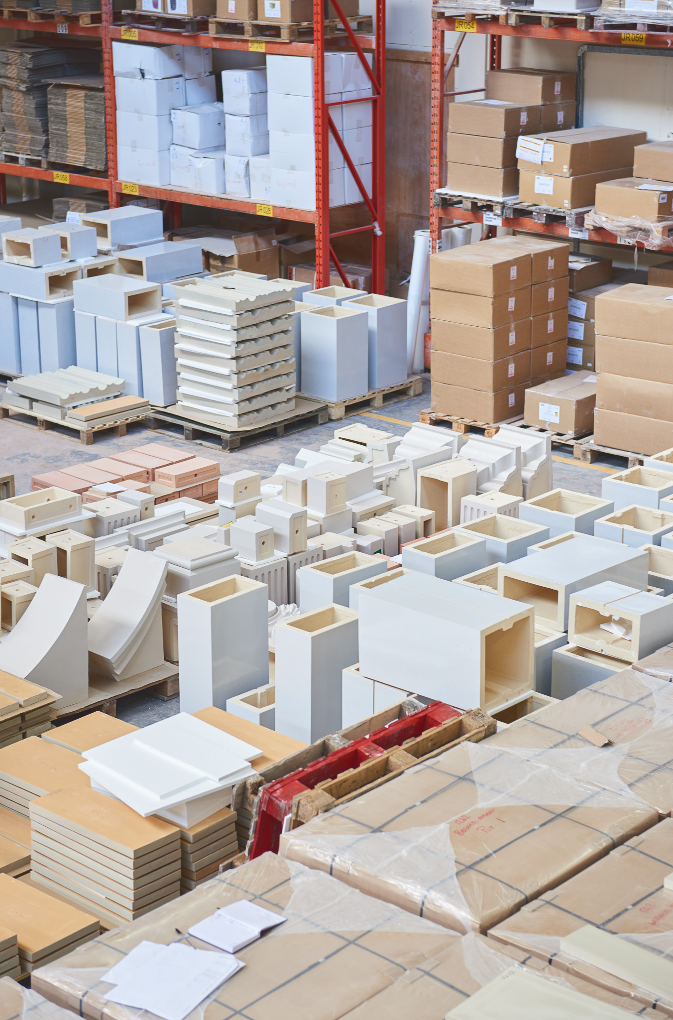 Stacks of ceramic sinks and cardboard boxes are arranged in a warehouse with shelves full of additional boxes and packages in the background.